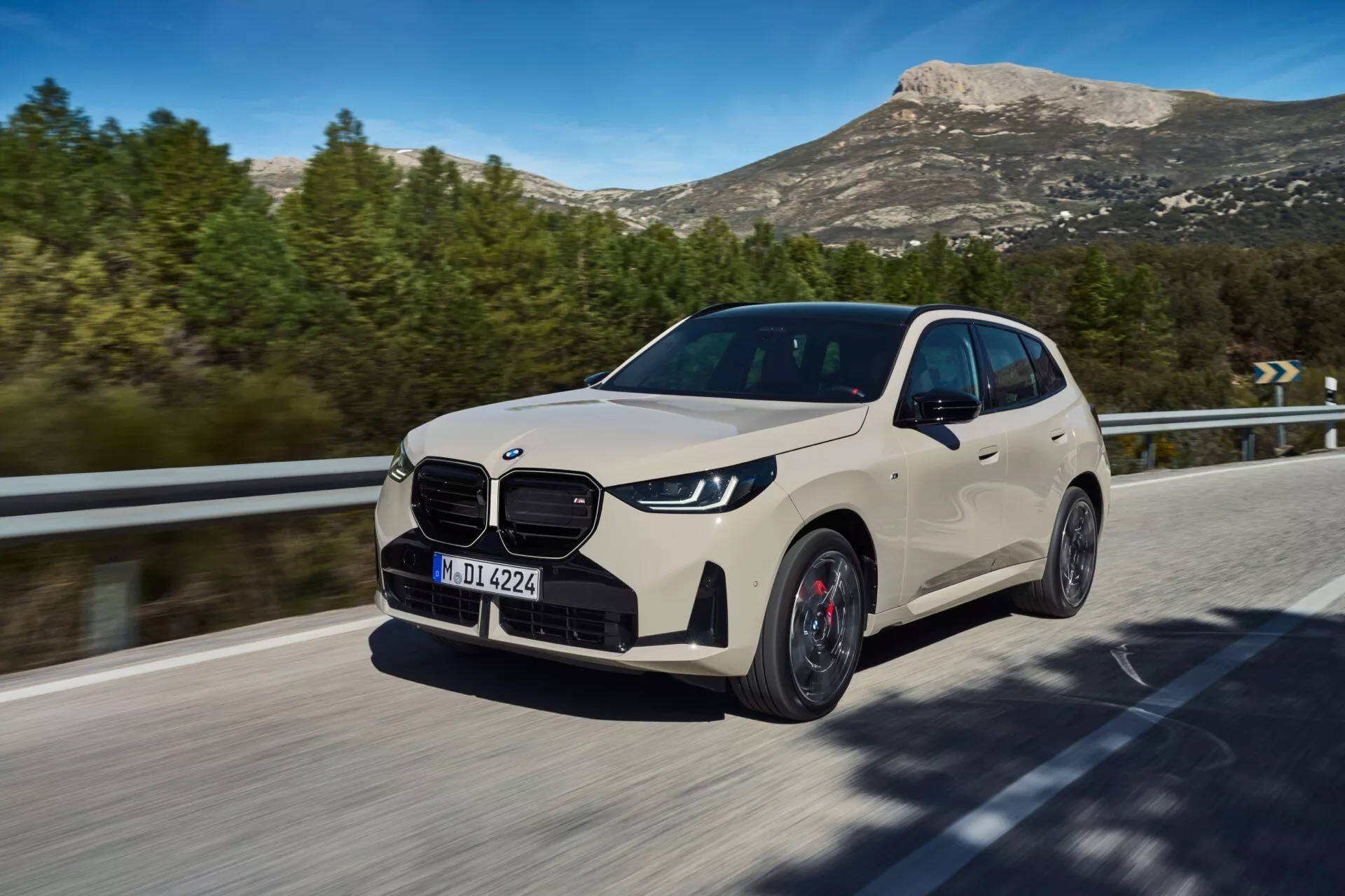 Dynamiczny kadr luksusowego crossovera BMW X3 2024 (G45) w ruchu na drodze, uchwycony z nieco niższego punktu widzenia. Ten trójwymiarowy przedni widok ukazuje charakterystyczną kratkę nerkową, nowoczesne kątowe światła do jazdy dziennej LED, przedni zderzak i część maski. Widoczna jest bardziej strona kierowcy z projektem felg aluminiowych, liniami konturowymi nadwozia i lusterkami bocznymi. Rozmyte tło i obracające się przednie koło sugerują ruch, z naturalnym górzystym krajobrazem w tle, co wskazuje na solidność i wszechstronność. Naturalne światło podkreśla design samochodu, akcentując zaawansowaną technologię i elementy aerodynamiczne, odzwierciedlając połączenie luksusu, wydajności i oszczędności.