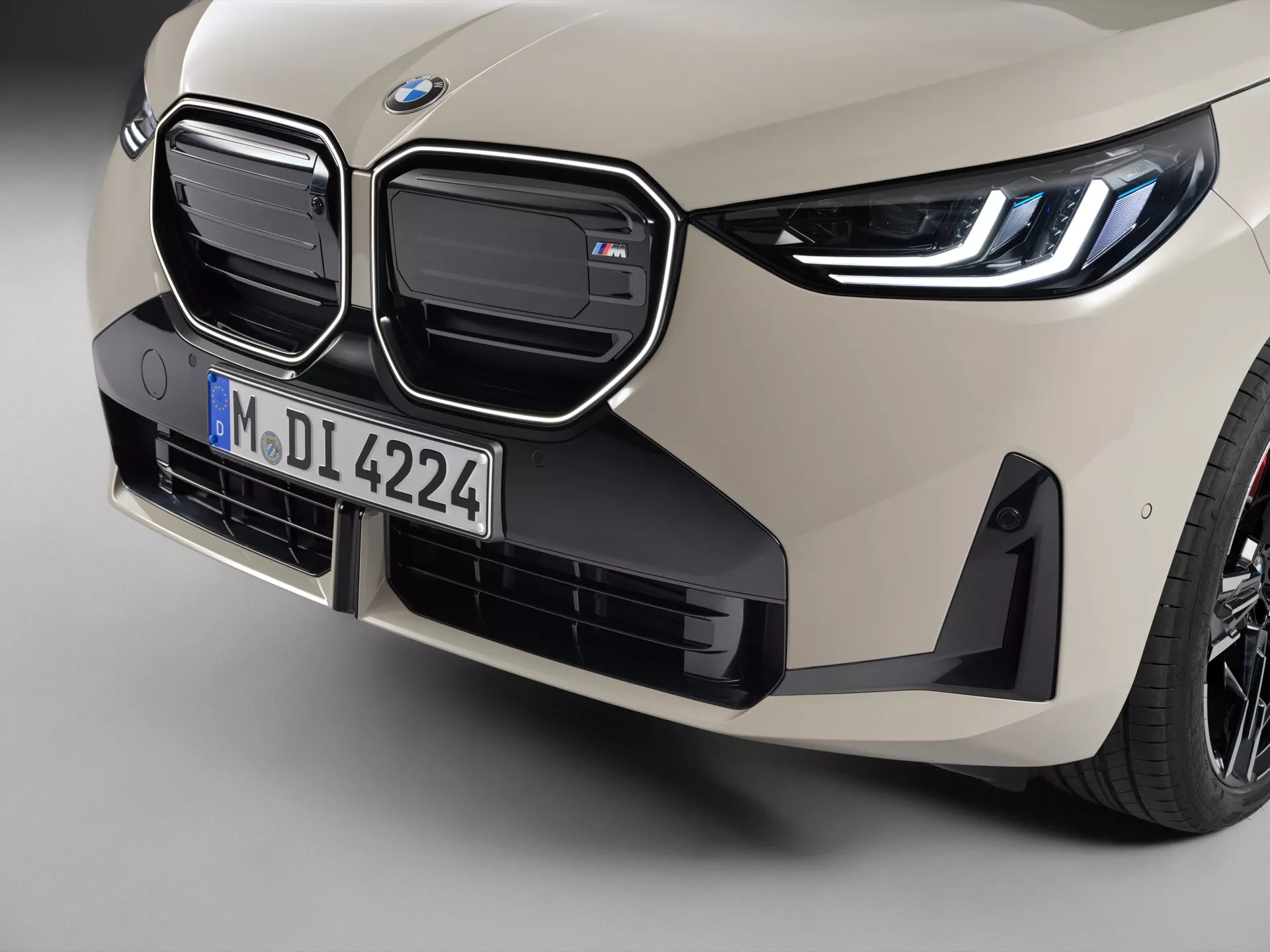 Zbliżenie na przednią lewą stronę BMW X3 (G45) 2024, prezentujące charakterystyczne podwójne nerki z chromowanymi obrysami i aktywnymi zasłonami, emblemat BMW na masce, subtelne linie konturowe na masce, kątowe wloty powietrza i przednią wargę na zderzaku, czujnik parkowania, smukłe reflektory LED i jasny, piaskowy beżowy kolor. Obraz podkreśla zaawansowany design i technologie pojazdu, koncentrując się na cechach aerodynamicznych i chłodzeniu, z płytką głębią ostrości akcentującą kratownicę oraz zderzak.