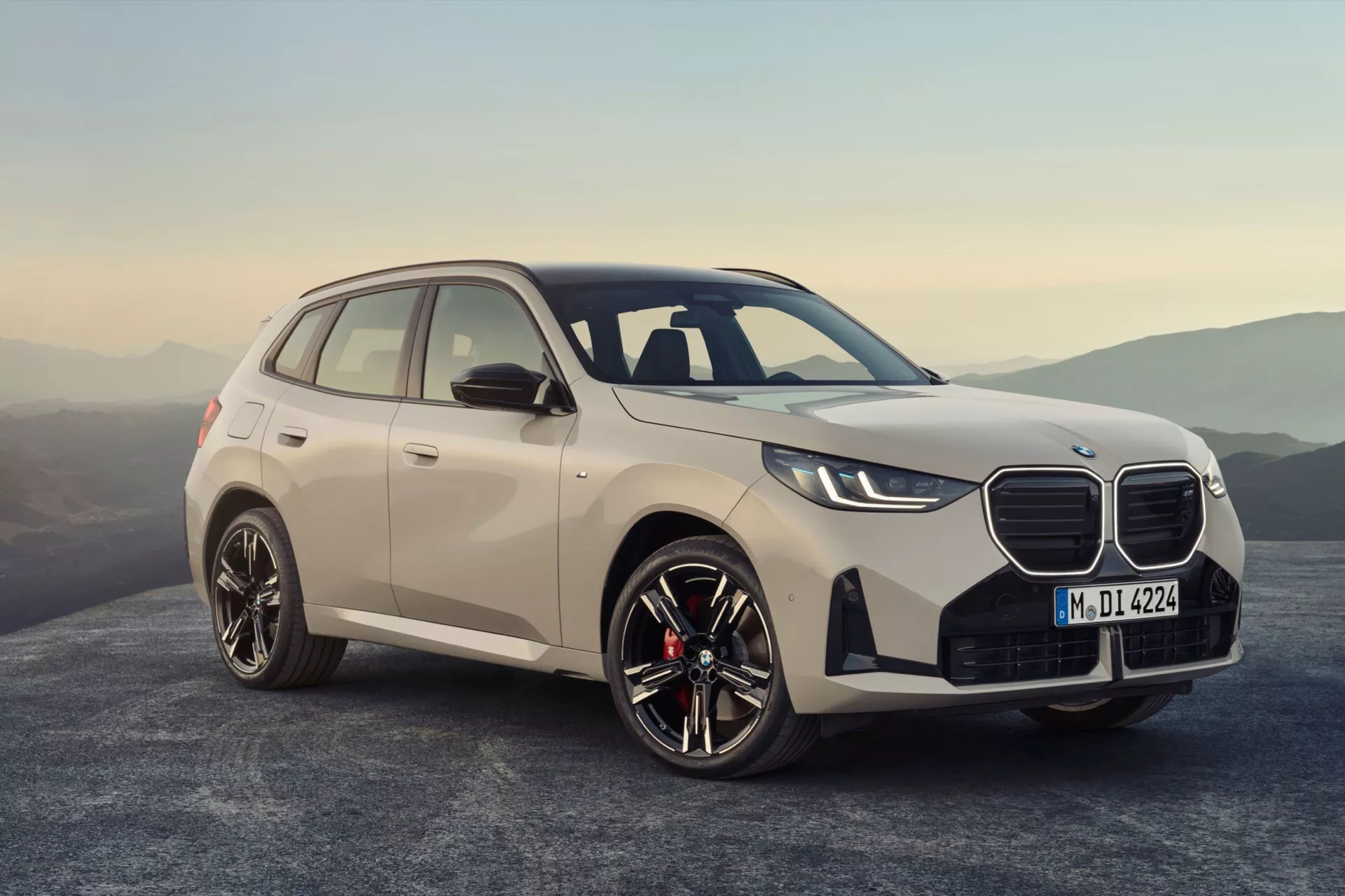 BMW X3 2024 (G45) widok z przodu z trzech czwartych, podkreślający przedni grill, smukłe reflektory LED, aerodynamiczne kontury maski, duże felgi aluminiowe i solidną sylwetkę SUV-a. Cechy obejmują lusterka boczne w kolorze nadwozia z kierunkowskazami, okna z chromowanymi ramkami i czerwone zaciski hamulca, sugerując orientację na osiągi. Ustawiony na tle gór o zmierzchu lub świcie, obraz podkreśla luksus, nowoczesny design i zaawansowaną technologię motoryzacyjną, w tym potencjalne systemy wspomagania i bezpieczeństwa.