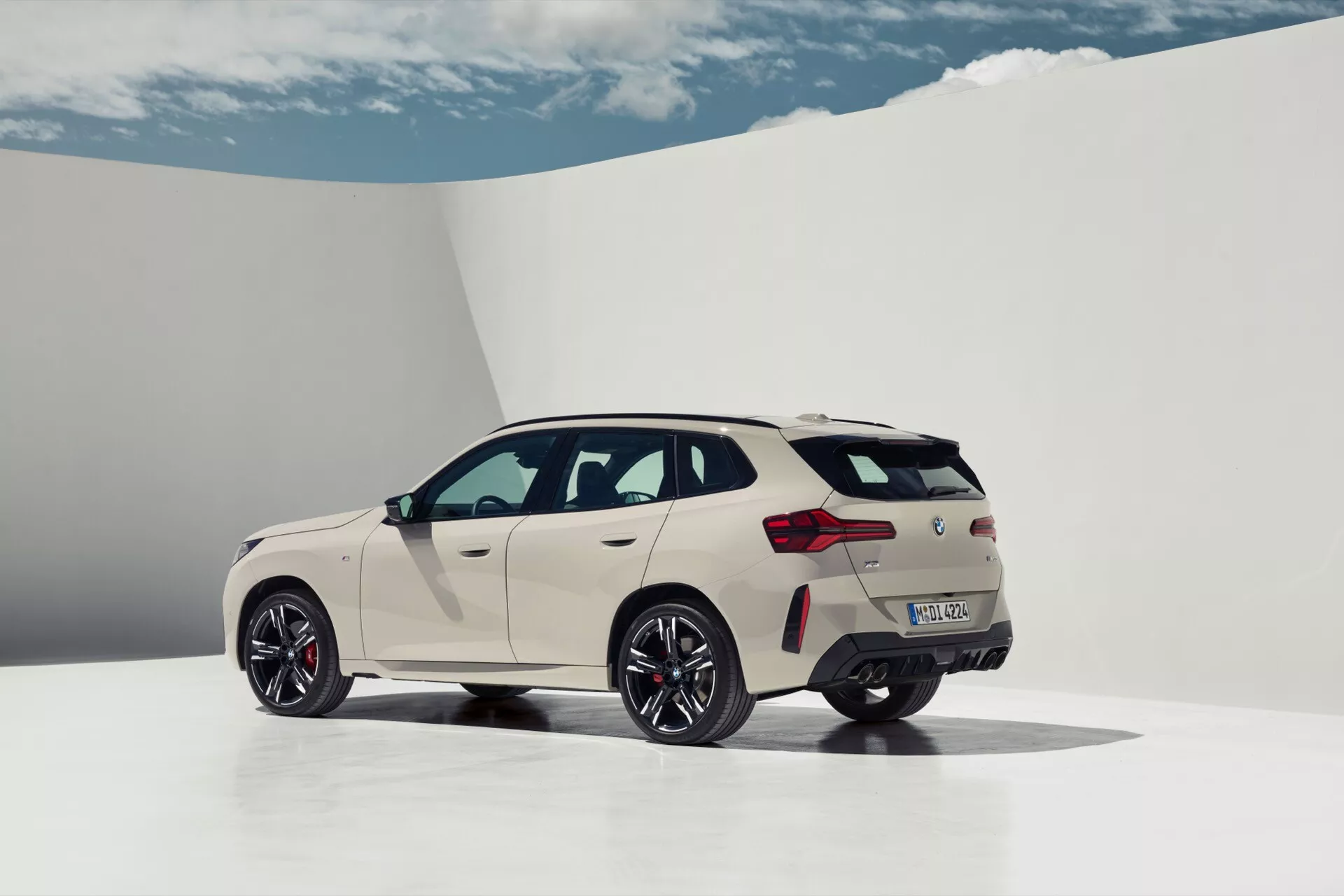 Zdjęcie przedstawia nowoczesne, sportowe SUV BMW X3 2024 (G45) z trzech czwartych tyłu, ukazując zarówno profil boczny, jak i tylną część pojazdu. Samochód pomalowany na jasny, prawdopodobnie perłowy biały lub bardzo jasny kremowy kolor, stoi w jasnym studio, które podkreśla jego linie i styl. Duże, ciemne wielo-ramienne koła i opony o niskim profilu podkreślają sportowy charakter pojazdu. Widać spojler na dachu, podwójne końcówki wydechu, poziome tylne światła i eleganckie linie charakteru. Tylny emblemat jest nieczytelny, tylne okna wydają się przyciemniane, obecne są lusterka boczne w kolorze nadwozia oraz antena typu płetwa rekina na dachu.