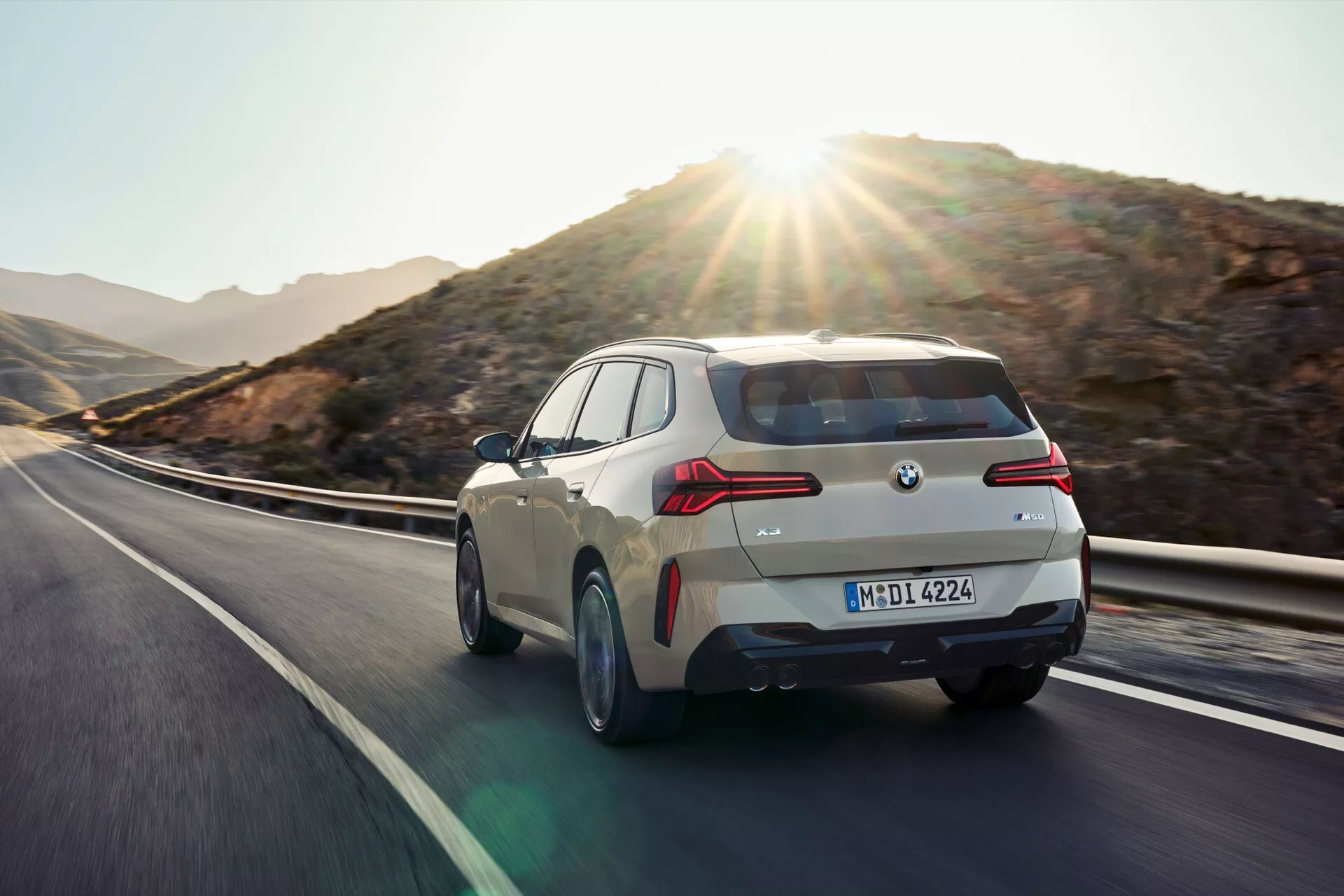 Obraz BMW X3 2024 (G45) zrobiony pod kątem trzy czwarte od tyłu, pokazujący tył i częściowy profil boczny pojazdu w ruchu. Podkreśla tylny design, w tym tylne lampy, tylny zderzak i krzywiznę samochodu. Inne widoczne cechy to tablica rejestracyjna i odznaka, tylna szyba, spoiler dachowy i antena w kształcie płetwy rekina. Jasny, błyszczący lakier samochodu odbija światło otoczenia, z naturalnym krajobrazem i jasnym słońcem w tle. Sugerowane są funkcje technologiczne takie jak tylne światła LED lub laserowe i aerodynamiczne projekty.