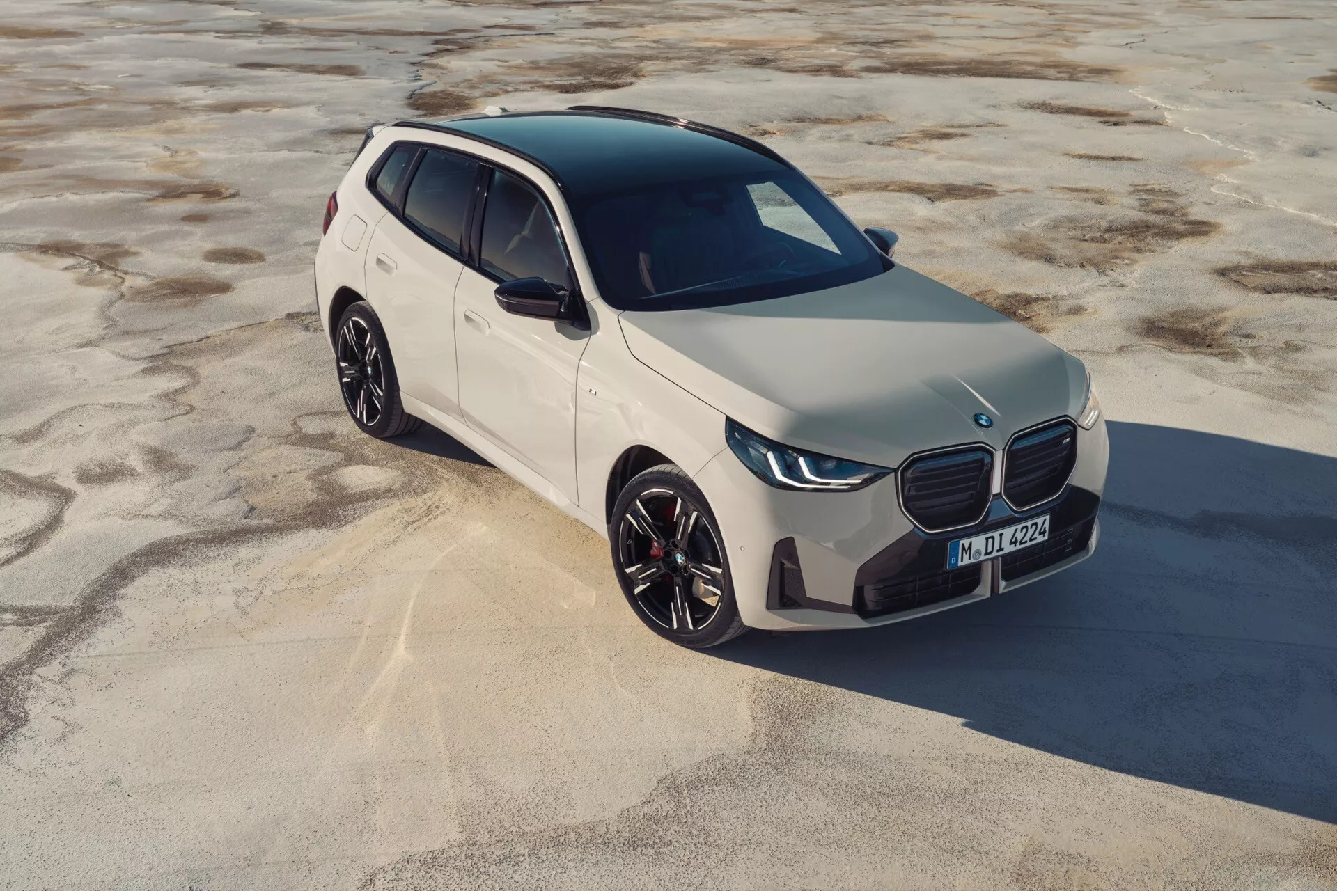 Widok z lotu ptaka pod kątem frontowo-trzyczwartowym na nowoczesnego kompaktowego SUV-a, BMW X3 2024 (G45). Pojazd stoi na suchej, teksturowanej powierzchni, być może na piasku, pod naturalnym światłem słonecznym. Cechy obejmują eleganckie linie, wyraźny zamknięty przedni grill, stylizowane reflektory LED, duże wieloramienne felgi aluminiowe i nachyloną przednią szybę wskazującą na aerodynamiczny profil. Kolor samochodu jest jasny, prawdopodobnie biały. Zamknięty grill sugeruje napęd elektryczny lub hybrydowy.