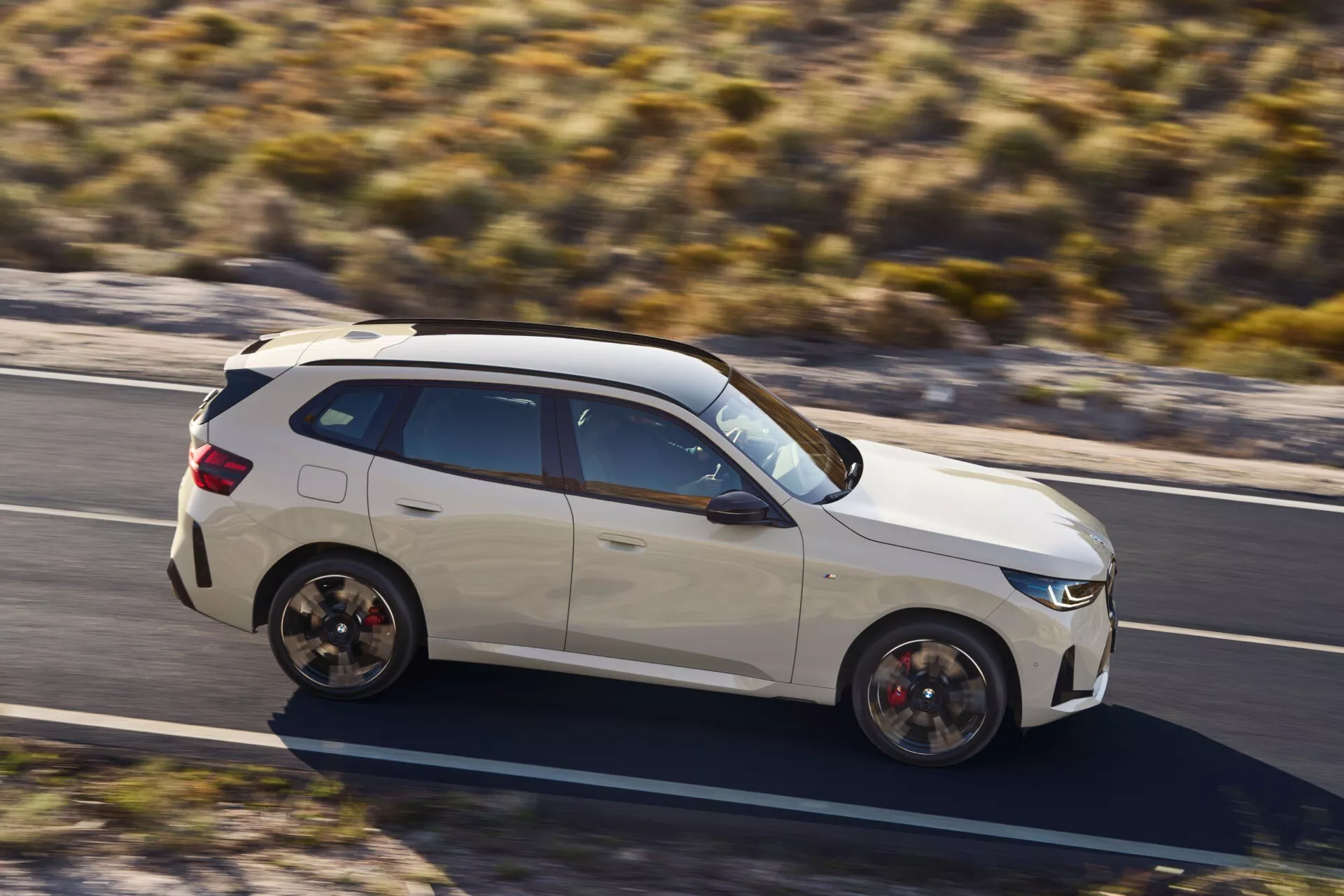 Widok z dużej wysokości na biały, sportowy SUV BMW X3 2024 (G45) w ruchu w pogodny dzień, prezentujący szczegółowy profil boczny z czarnymi elementami, pochylonym dachem i dużymi felgami z widocznymi zaciskami hamulcowymi, sugerujące wysoką wydajność i zaawansowane funkcje technologiczne.