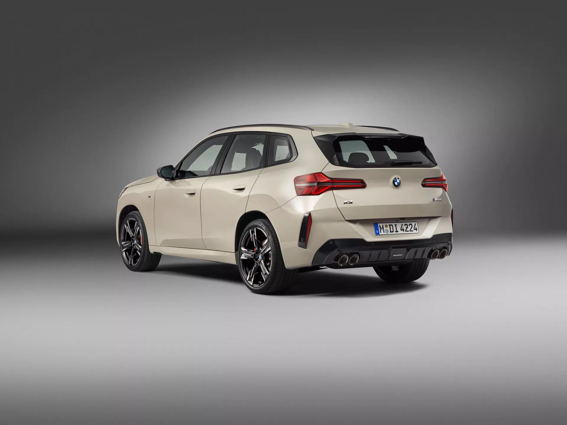 Widok tylno-trzyczwartkowy nowoczesnego hatchbacka BMW, uchwycony z niskiego kąta, przedstawiający posturę i linie projektowe pojazdu. Jasny kremowy lub biały kolor kontrastuje z czarnymi wykończeniami wokół okien, dolnej części karoserii oraz ciemnymi, wieloramiennymi felgami aluminiowymi. Tylne elementy obejmują tylne światła LED owinięte wokół boków, podwójny układ wydechowy, dyfuzor, antenę w kształcie płetwy rekina i tylne wycieraczki. Widoczne detale technologiczne obejmują czujniki parkowania na zderzaku, sugerujące system asystenta parkowania, oraz możliwość kamery cofania pod emblematem. Neutralne szare tło uwydatnia samochód, z miękkim, rozproszonym oświetleniem zapewniającym wygląd premium.