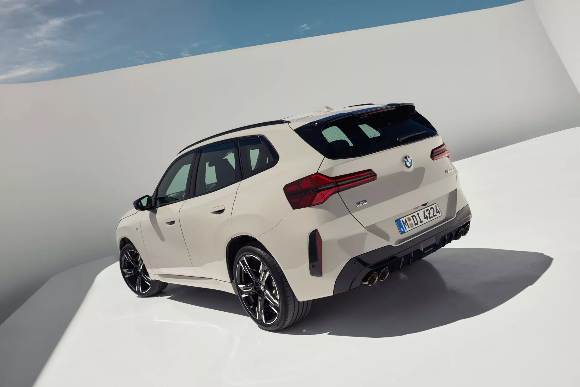 Widok z wysokiego trzech czwartych tyłu nowoczesnego luksusowego SUV-a BMW X3 2024 (G45). Obraz podkreśla sportowy wygląd i estetykę projektowania, w tym tylne światła LED o kanciastym kształcie, tylny spojler, duże, wieloszprychowe obręcze aluminiowe z oponami wysokiej wydajności i duże tarcze hamulcowe. Charakterystyczne linie i rzeźbiona powierzchnia potęgują dynamiczny wygląd, podczas gdy zlicowane klamki drzwi sugerują nowoczesną technologię. Perłowobiała farba uwydatnia krzywizny, a zarys panoramicznego dachu sugeruje luksusowe wnętrze. Czyste tło zapewnia, że uwaga skupia się na wyrafinowanym wzornictwie i zaawansowanych funkcjach pojazdu.