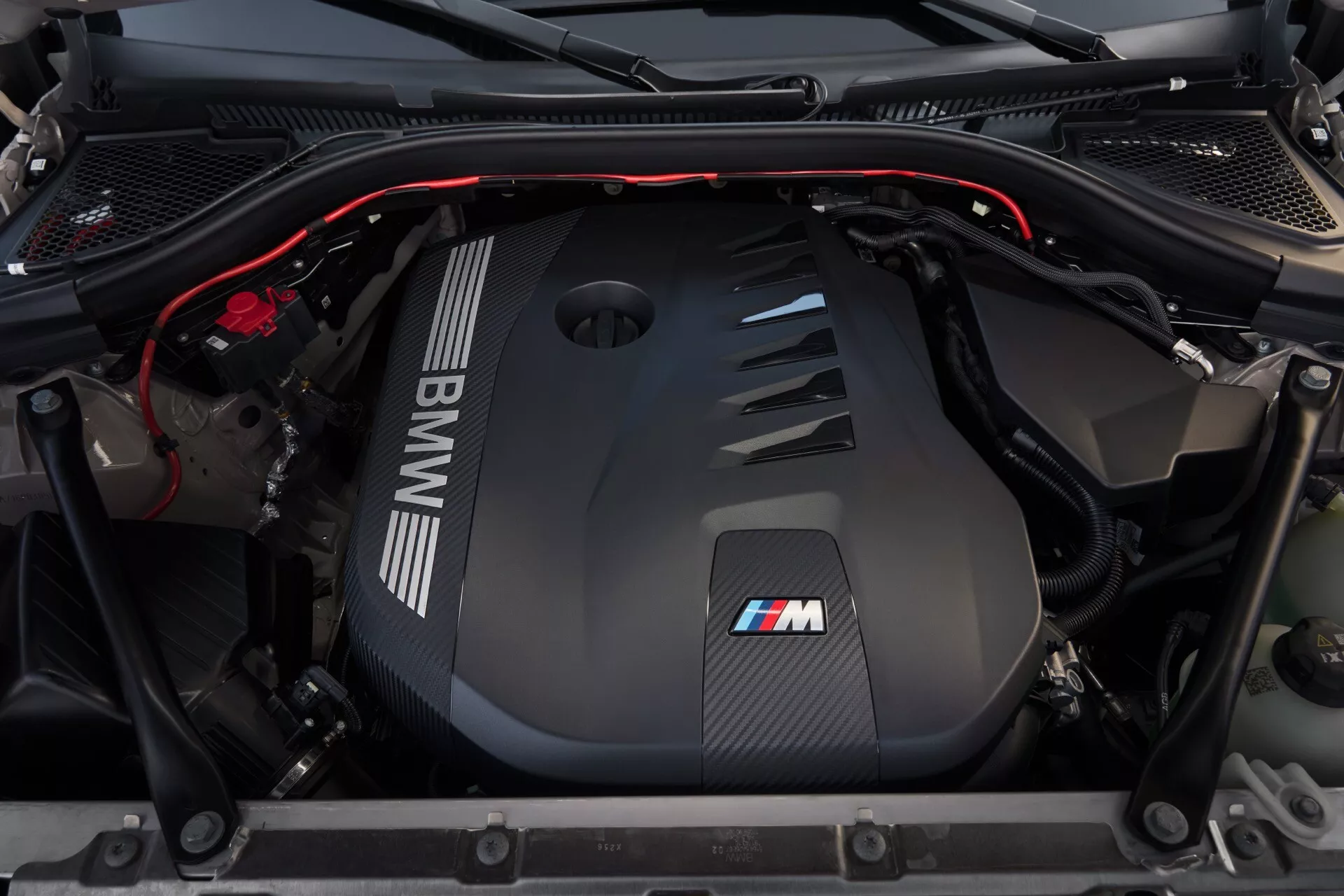 Widok z góry i przodu komory silnika BMW X3 2024 (G45), prezentujący pokrywę silnika z włókna węglowego z wyraźnym logo BMW i odznaką M. Szczegółowy zbliżenie podkreśla lekkie materiały, kolektor ssący, belkę wzmacniającą i czerwone linie akcentujące, oznaczające elementy wysokowydajnego designu.