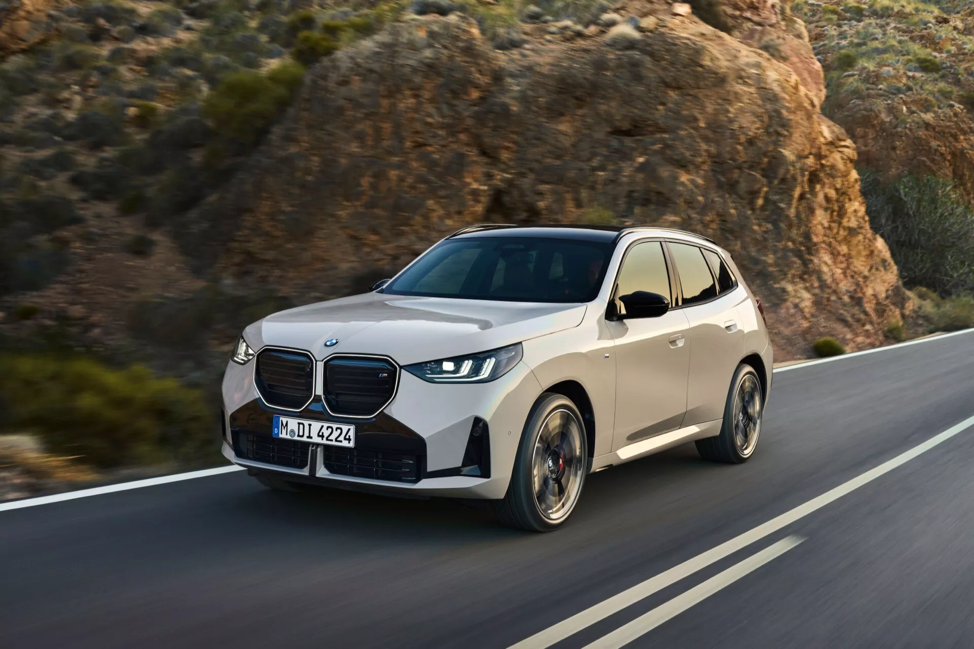 Obraz BMW X3 2024 (G45) w ruchu na krętej drodze, z skalistym krajobrazem w tle. Zdjęcie, wykonane pod kątem przedniego ćwierć, uwydatnia lewą przednią stronę pojazdu, pokazując szczegółowe cechy jak duża przednia krata, agresywne reflektory LED, wloty powietrza, konturowana maska, aerodynamiczne lusterka boczne, nowoczesne felgi aluminiowe oraz lekko przyciemniane szyby. Elementy designu i technologii samochodu, wskazujące na funkcje bezpieczeństwa i wydajności, są wyraźnie widoczne.