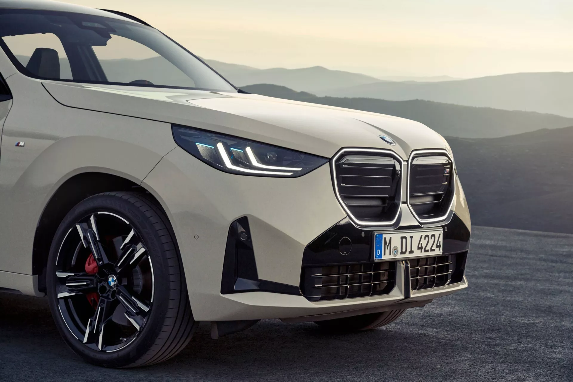 Zbliżenie pod niskim kątem na lewy przedni róg BMW X3 2024 (G45), podkreślające charakterystyczną, nerkowatą osłonę chłodnicy z aktywnymi żaluzjami, wąskie, wydłużone reflektory LED z wbudowanymi kierunkowskazami, agresywny przedni zderzak z dużymi wlotami powietrza, profilowaną maskę, sportowe linie błotnika, premiumową wieloszprychową czarną felgę z wykończeniem obrabianym i czerwonym zaciskiem hamulcowym oraz aerodynamiczne lusterko boczne z wbudowanym kierunkowskazem; na rozmytym górskim tle.