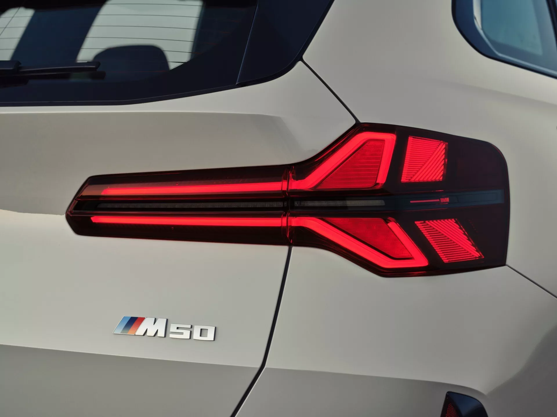Zbliżenie tylnej części BMW X3 2024 (G45), skupiając się na projekcie tylnego światła z ostrymi, kanciastymi liniami i iluminowaną technologią LED. Tylne światło posiada kombinację przezroczystych i czerwonych części, co wskazuje na system świateł do jazdy dziennej lub hamowania, a obok znajduje się oznaczenie 'M50', sugerujące model o wysokich osiągach. Karoseria samochodu ma zakrzywioną, lśniącą powierzchnię z perłowobiałym wykończeniem, uwieczniając szczegóły i refleksy w wysokiej rozdzielczości w naturalnym świetle dziennym.