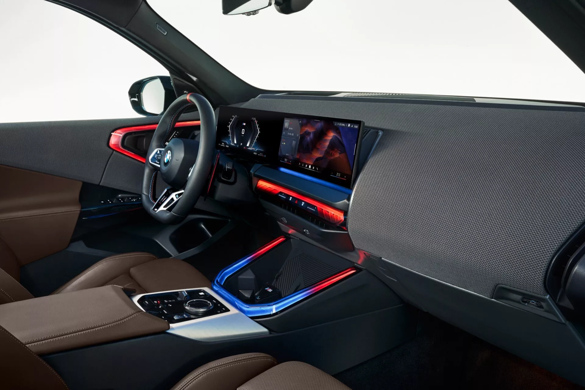 Widok wnętrza BMW X3 2024 (G45) z perspektywy pasażera, ukazujący przestrzeń kierowcy, elegancki kokpit z dużym wyświetlaczem dotykowym, niebieskie oświetlenie ambientowe, premium dwukolorowe fotele skórzane i panele drzwi, elektroniczny selektor biegów otoczony dotykowymi kontrolkami oraz nowoczesną, luksusową estetykę.