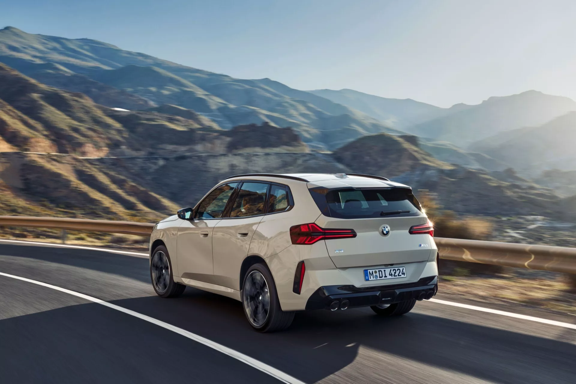Dynamiczny widok z trzech czwartych tyłu nowoczesnego hatchbacka BMW X3 2024 (G45) uchwycony w ruchu na górskiej drodze. Obraz podkreśla sportowy design z liniami charakteru, zintegrowanym tylnym spojlerem, widocznymi podwójnymi wylotami wydechu i nowoczesnymi tylnymi światłami LED. Samochód znajduje się na prawym pasie, z falującymi wzgórzami i czystym niebem w tle. Rozmazane linie na drodze i krajobrazie sugerują prędkość, podkreślając charakter pojazdu nastawiony na osiągi.