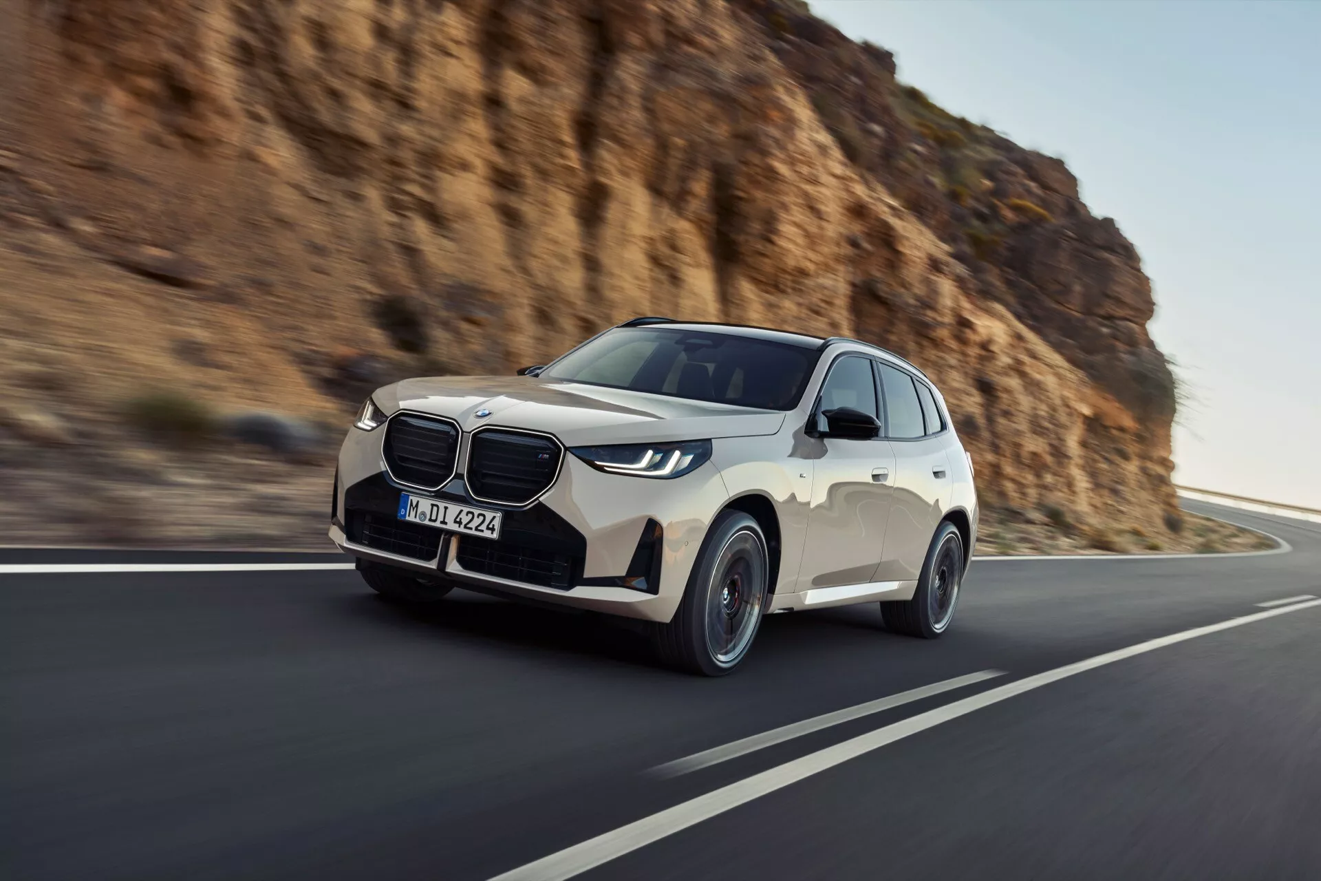 Dynamiczne ujęcie ruchu BMW X3 2024 (G45) wykonane pod niskim, frontowym kątem, podkreślające ruch i moc. Cechy obejmują wyróżniającą się, zamkniętą kratkę z chromowanymi akcentami, eleganckie kątowe reflektory LED, dwukolorowy wygląd zewnętrzny z czarnym dolnym wykończeniem, aluminiowe felgi o wieloramiennym wzorze oraz lusterka boczne w kolorze nadwozia z zintegrowanymi kierunkowskazami. Samochód znajduje się po prawej stronie nadmorskiej lub górskiej drogi z rozmytym tłem akcentującym prędkość, uchwycony w naturalnym świetle.