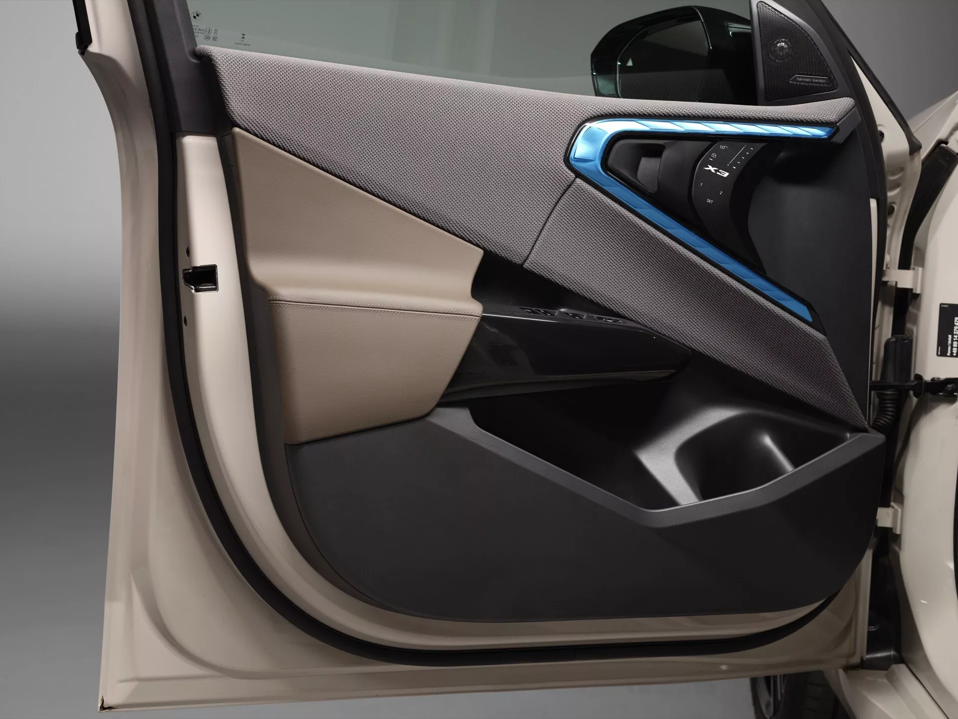 Zbliżenie na wewnętrzny panel drzwi samochodu BMW X3 2024 (G45) uchwycony pod kątem ukośnym. Widać ergonomiczną klamkę z niebieskim oświetleniem ambientowym, zintegrowane kieszenie drzwiowe, jasne wykończenie kontrastujące z ciemnymi plastikowymi materiałami, bezszwowy design, panele sterowania potencjalnymi regulacjami okien i lusterek oraz nowoczesne, luksusowe dwukolorowe schematy kolorystyczne.