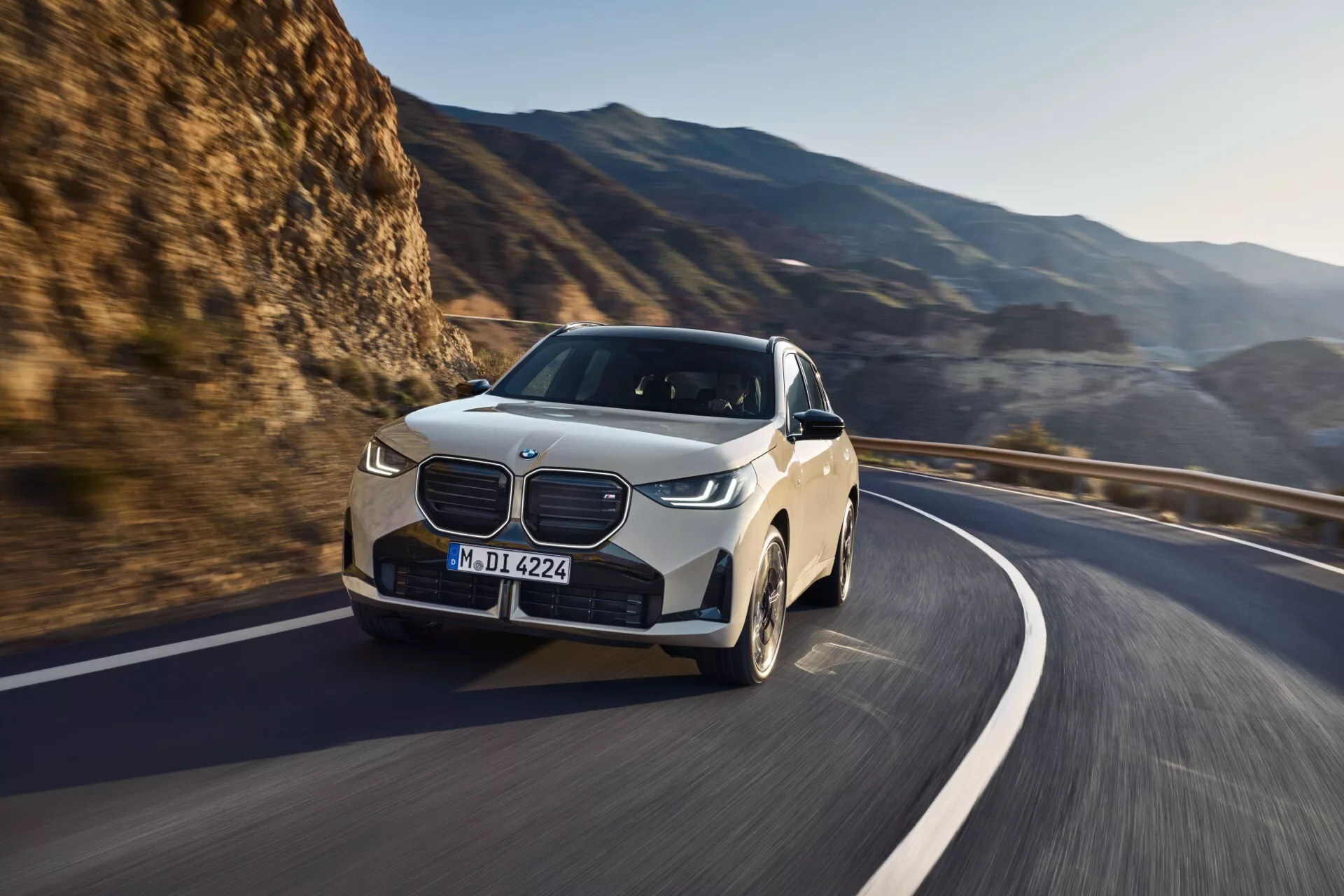 BMW X3 2024 (G45) uchwycone w ruchu z przedniego widoku pod kątem trzech czwartych pokazuje nowoczesny design SUV-a, z wyraźnymi cechami takimi jak duży wyraźny grill, cienkie przednie światła LED, zaokrąglona maska i duże aluminiowe felgi. Uchwycone podczas jazdy na zakrzywionej górskiej drodze przy jasnym świetle słonecznym, zdjęcie podkreśla sportowy, aerodynamiczny styl i luksusowy wygląd pojazdu. Scena sugeruje zaawansowane możliwości jazdy i technologie takie jak kontrola stabilności i adaptacyjne reflektory, jednak wewnętrzne cechy i specyficzne szczegóły techniczne nie są widoczne.