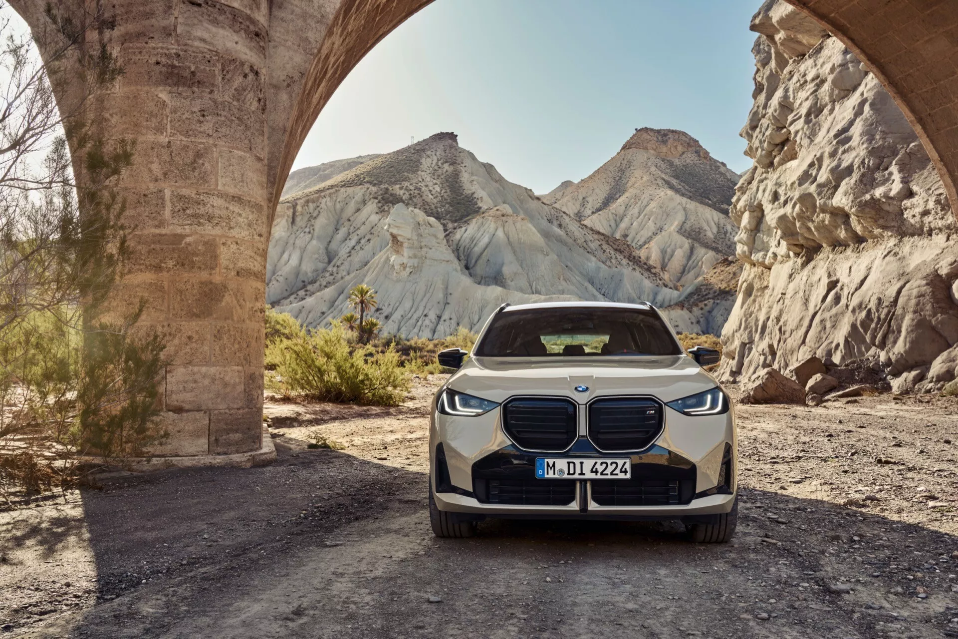 Fotografia BMW X3 2024 (G45) z kąta przedniego, podkreślająca charakterystyczną nerkowatą kraty, eleganckie reflektory LED z dziennymi światłami i przedni zderzak z centralnym i bocznymi wlotami. Pojazd jest ustawiony centralnie w rustykalnym otoczeniu na świeżym powietrzu pod kamiennym łukiem z jałowymi wzgórzami w tle, pokazując potencjalne zdolności terenowe i zaawansowane technologie motoryzacyjne.