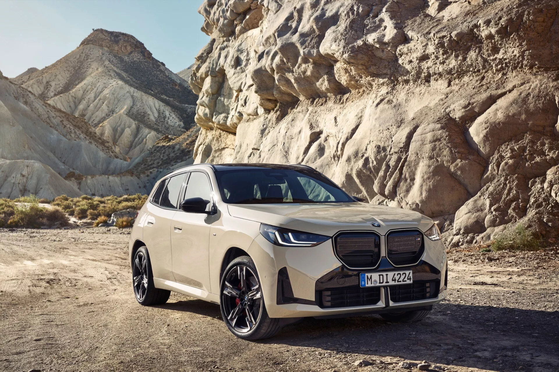 Widok z trzech czwartych przodu SUV-a BMW X3 (G45) 2024 roku, prezentujący jego nowoczesny i ostry design z wyraźnym podzielonym grillem z emblematem marki, eleganckimi reflektorami LED, rzeźbioną maską i wyrafinowanym przednim zderzakiem z wlotami powietrza. Profil kierowcy ujawnia duże felgi aluminiowe, wyraźne linie boczne, zintegrowane lusterka i panoramiczny dach. Umieszczony na suchej, skalistej powierzchni z pustynnym tłem, pojazd w jasnym neutralnym kolorze podkreśla luksus, zaawansowaną technologię i wszechstronne zdolności zarówno na drodze, jak i poza nią.
