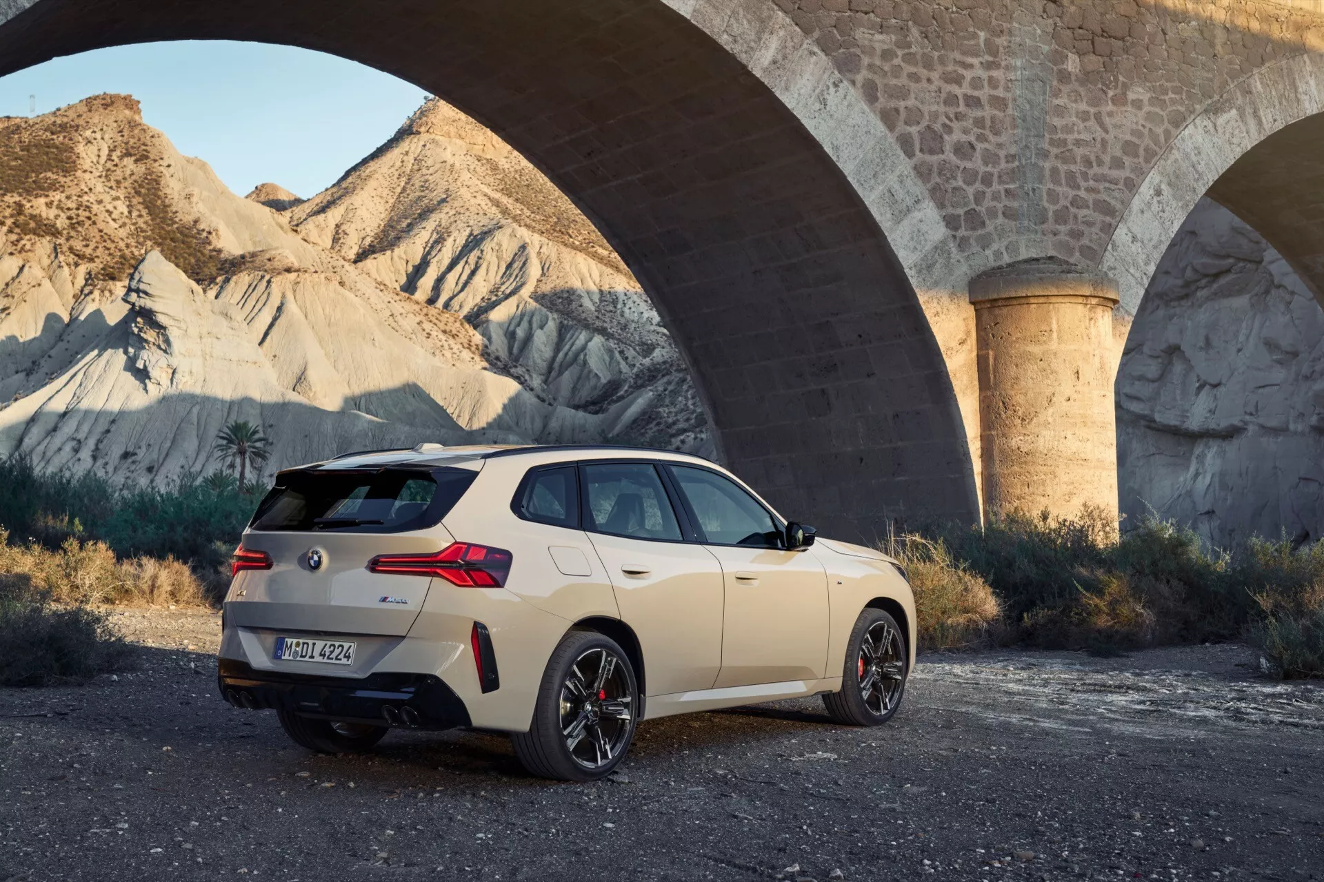 Obraz przedstawia hatchback BMW X3 2024 (G45), zaparkowany na nieutwardzonej powierzchni, widziany pod kątem tylno-trzeciowym. Samochód posiada sportowe felgi aluminiowe, spojler dachowy, tylne okno z wycieraczką, opakowane diodowe światła tylne, podwójne wyloty wydechu i emblematy z tyłu. Stoi pod dużym betonowym mostem w scenerii późnego popołudnia, a jego kremowobiały kolor kontrastuje z ziemistym tłem.