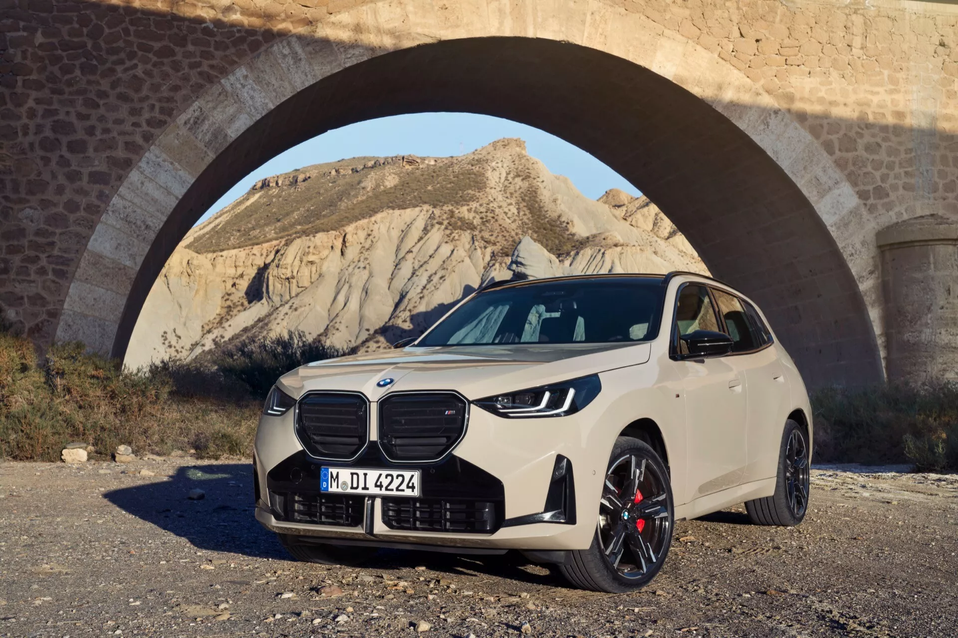 Widok z przodu w trzy czwarte BMW X3 (G45) 2024, z wyeksponowaną powiększoną nerkowaną kratką z pionowymi listwami, eleganckimi reflektorami LED lub potencjalnie laserowymi, przednią kamerą, sportowymi wieloramiennymi felgami z czerwonymi zaciskami hamulcowymi, co wskazuje na wysokowydajne hamulce, oraz ewentualnie pakietem M Sport z dodatkowymi wlotami powietrza lub światłami przeciwmgielnymi na przednim zderzaku. Samochód w kremowym lub jasnobeżowym odcieniu jest ustawiony pod łukiem w naturalnym świetle dziennym, tworząc malownicze otoczenie, które podkreśla zarówno elementy architektoniczne, jak i naturalne krajobrazy, podnosząc jego nowoczesny design i zaawansowane funkcje.