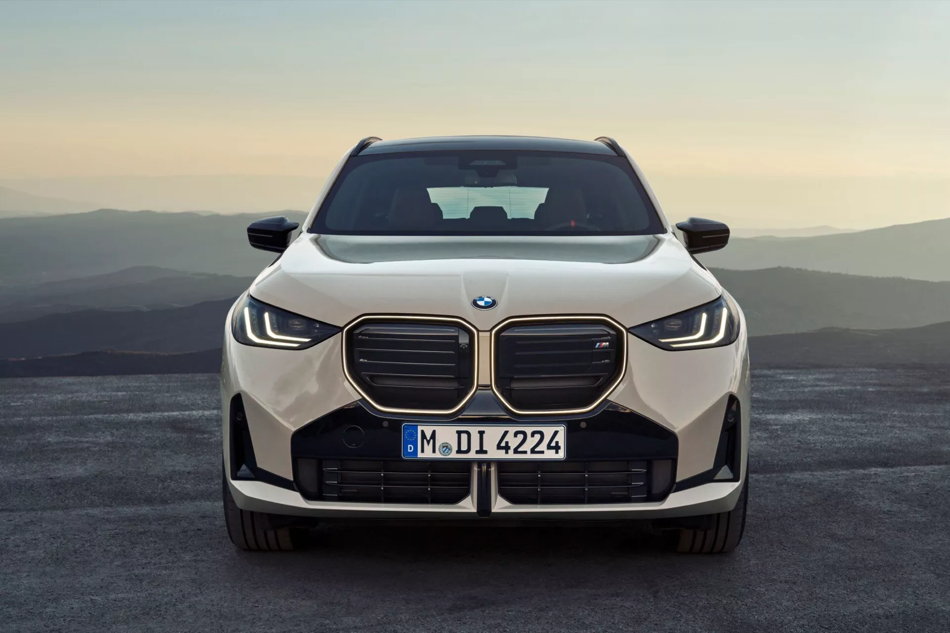 Widok z przodu BMW X3 2024 (G45) uchwycony na poziomie oczu, z charakterystycznym grillem przypominającym nerki otoczonym eleganckimi reflektorami LED o kanciastym kształcie, sugerującymi adaptacyjne technologie reflektorów. Samochód jest ustawiony na tle rozległego pleneru w ciepłym świetle wschodu lub zachodu słońca. Centralny wlot powietrza i sportowy, a zarazem elegancki przedni zderzak z widocznymi czujnikami parkowania sugerują zaawansowane systemy wspomagania kierowcy. Maska samochodu ma gładkie linie i subtelne zagięcia, które poprawiają profil aerodynamiczny, z wyraźnie widocznym emblematem BMW na końcu.