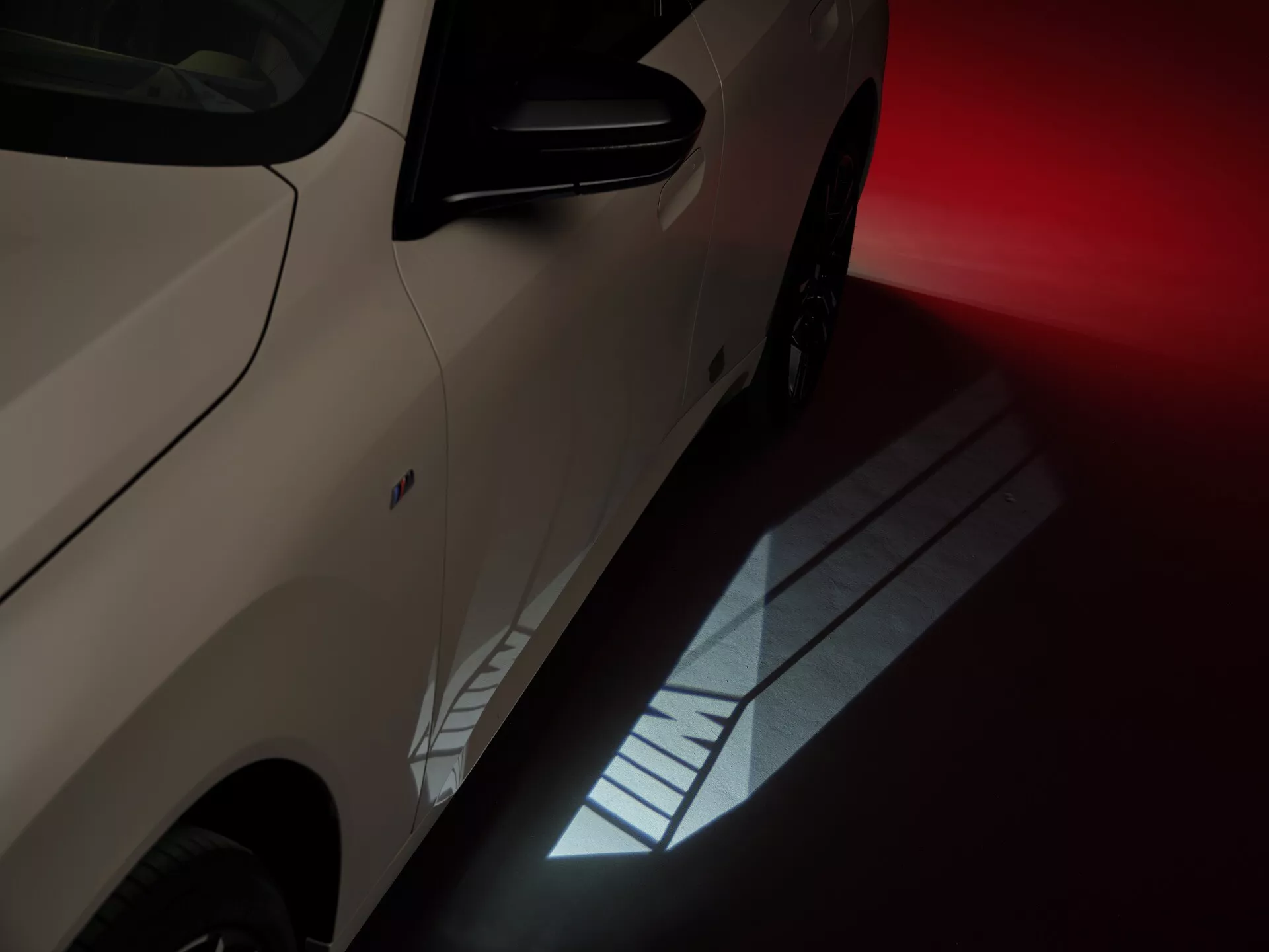 Zbliżenie i kątowe zdjęcie bocznego profilu BMW X3 2024 (G45), pokazujące przednie koło, błotnik i część przednich drzwi pasażera z widoczną felgą aluminiową i klamką drzwi. Oświetlenie tworzy dramatyczną atmosferę z silnymi odbiciami na metalicznym, szampańskim lakierze samochodu, sugerując ustawienie w salonie lub stylizowanej sesji zdjęciowej.