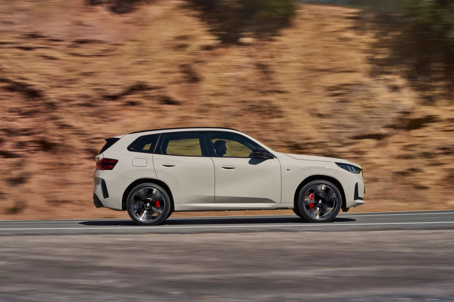 BMW X3 2024 (G45) w ruchu, uchwycone na zdjęciu panoramującym, aby podkreślić prędkość i dynamiczną postawę. Profil boczny pokazuje eleganckie linie projektowe, cztery drzwi, duże aluminiowe felgi z czerwonymi zaciskami hamulcowymi, okna w chromowanych ramach i szyberdach. Tło jest rozmyte, gdy samochód przemieszcza się przez suchy, pagórkowaty krajobraz.
