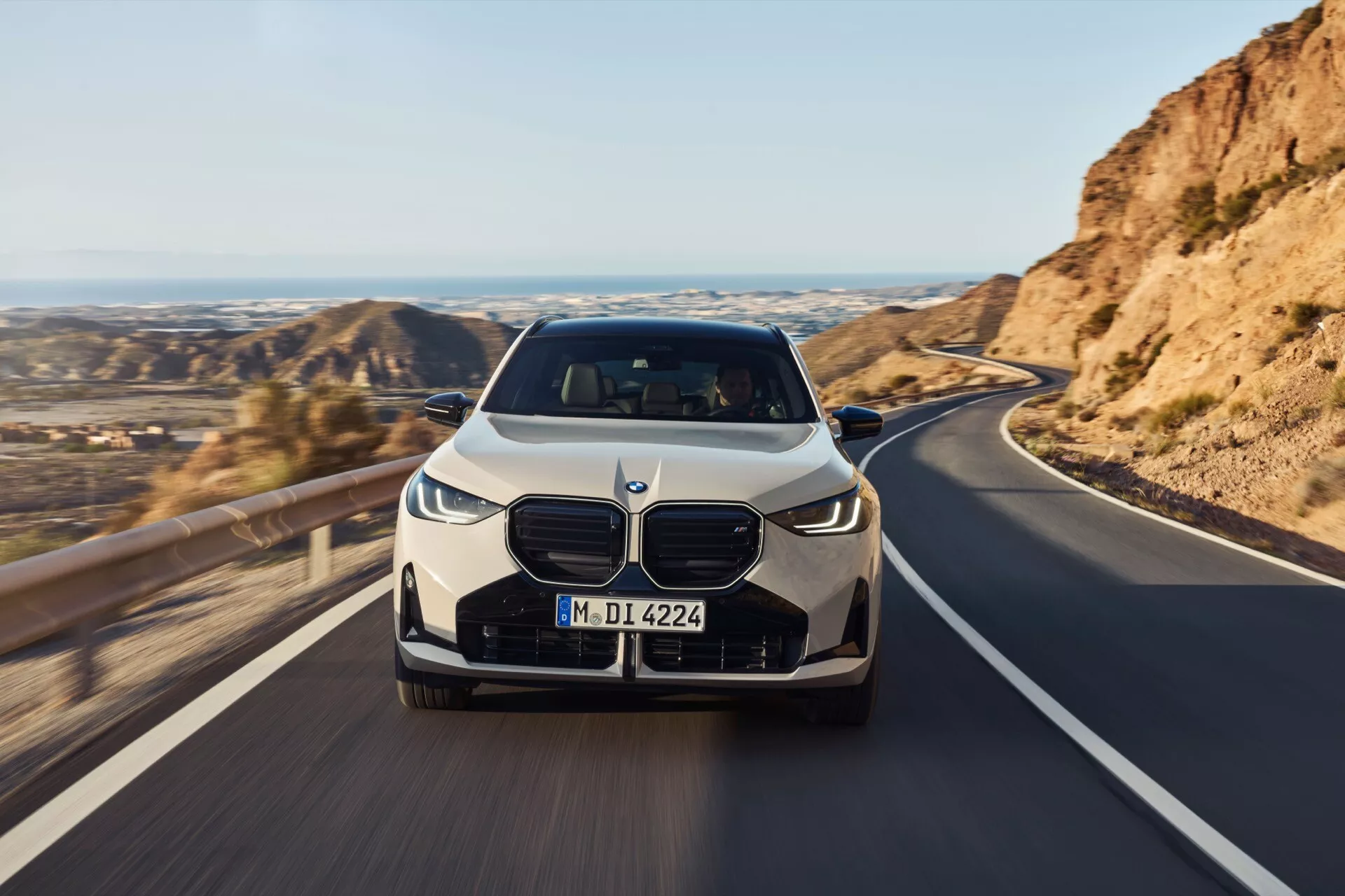 Widok z przodu BMW X3 2024 (G45) na drodze z malowniczym górskim krajobrazem; cechy obejmują dużą kratkę, eleganckie reflektory LED, aerodynamiczny zderzak i linie konturowe na masce; możliwe zaawansowane technologie takie jak adaptacyjne LED, systemy wspomagania kierowcy i system kamer 360 stopni; obraz sugeruje ruch.