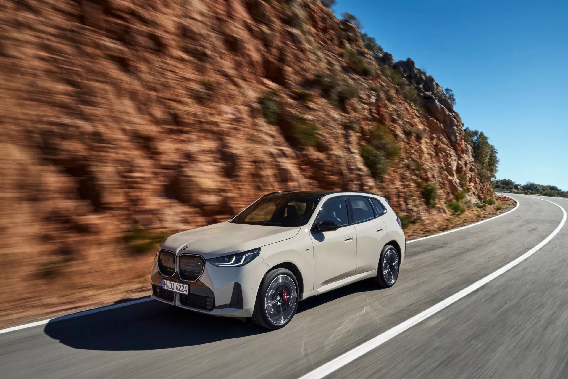 Dynamiczny obraz nowoczesnego SUV-a BMW X3 2024 (G45) uchwyconego w ruchu na otwartej, krętej drodze w ciągu dnia. Samochód widziany jest z perspektywy trzech czwartych z naciskiem na przednią lewą ćwiartkę, ukazując jego elegancki, sportowy design, wyrazisty przedni grill, ostre reflektory LED, duże sportowe koła i widoczne zaciski hamulcowe.