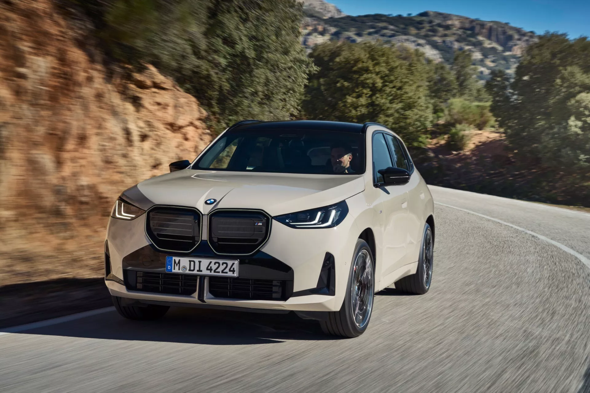 BMW X3 2024 (G45) w ruchu na krętej drodze z skalistym, wiejskim tłem. Widok z przodu pod kątem trzech czwartych uwydatnia charakterystyczny przedni grill z elementami w kształcie nerek, eleganckie reflektory LED, wieloramienne felgi aluminiowe oraz błyszczący jasno-neutralny kolor nadwozia. Widoczne są również aerodynamiczne lusterka boczne, konturowe linie nadwozia, przyciemniane szyby i centralny znaczek, podkreślając stylowy design i cechy premium.