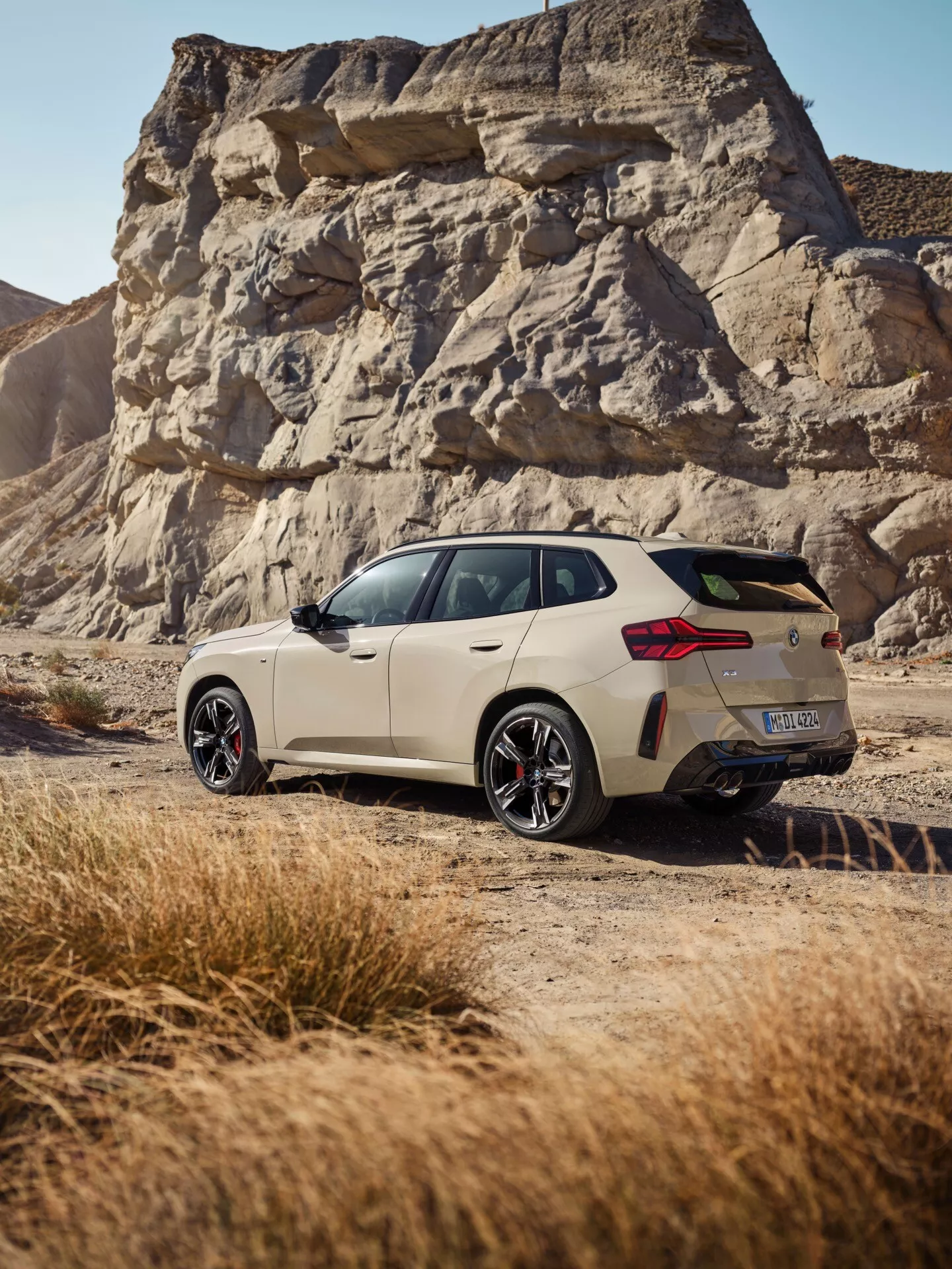 Obraz BMW X3 2024 (G45) uchwyca samochód z trzy-czwartego widoku tyłu na nieutwardzonej drodze pustynnej, pokazując jego charakterystyczne tylne elementy, w tym tylne światła LED, aerodynamiczny tylny spojler, sportowe wyloty wydechowe i felgi aluminiowe. Samochód w jasnym, matowym kolorze odbija popołudniowe światło słoneczne, z kamiennym krajobrazem w tle, sugerującym przygodę.
