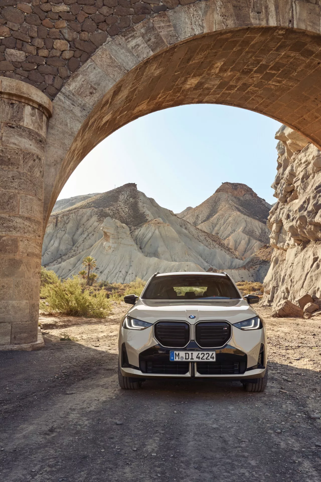 BMW X3 2024 (G45) widok z przodu fotografowany pod łukowatym kamiennym mostem. Samochód, umieszczony centralnie i zajmujący dolną połowę obrazu, pokazuje nowoczesne reflektory, charakterystyczny grill w kształcie nerek i emblemat BMW, co sugeruje model elektryczny lub hybrydowy plug-in. Sceneria pustynna z erodowanymi wzgórzami kontrastuje z zaawansowanym designem pojazdu, eksponując połączenie luksusu i nowoczesnej technologii motoryzacyjnej.