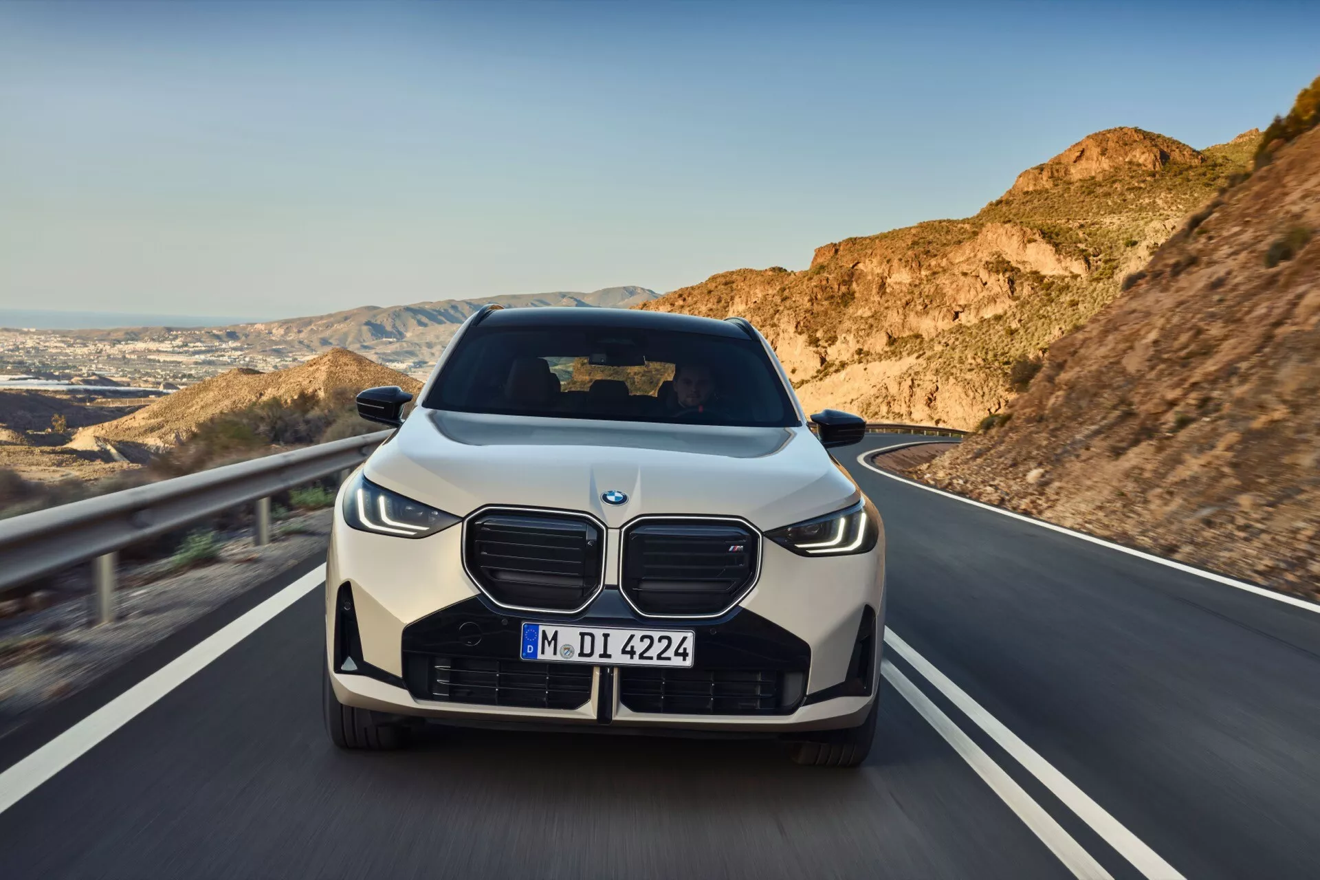 Widok z przodu na BMW X3 2024 (G45) ze wspaniałym grillem, zderzakiem, reflektorami i maską silnika. Samochód jedzie po krętej drodze z górami i czystym niebem w tle, co sugeruje ruch. Reflektory wyglądają na LED z dziennymi światłami, a duży grill sugeruje znaczące zapotrzebowanie na chłodzenie. Obraz sugeruje również zaawansowane systemy wspomagania kierowcy (ADAS), choć sensory nie są wyraźnie widoczne. Samochód przedstawiony jest w standardowym stanie fabrycznym, uchwycony wysokiej jakości aparatem, który podkreśla pojazd.