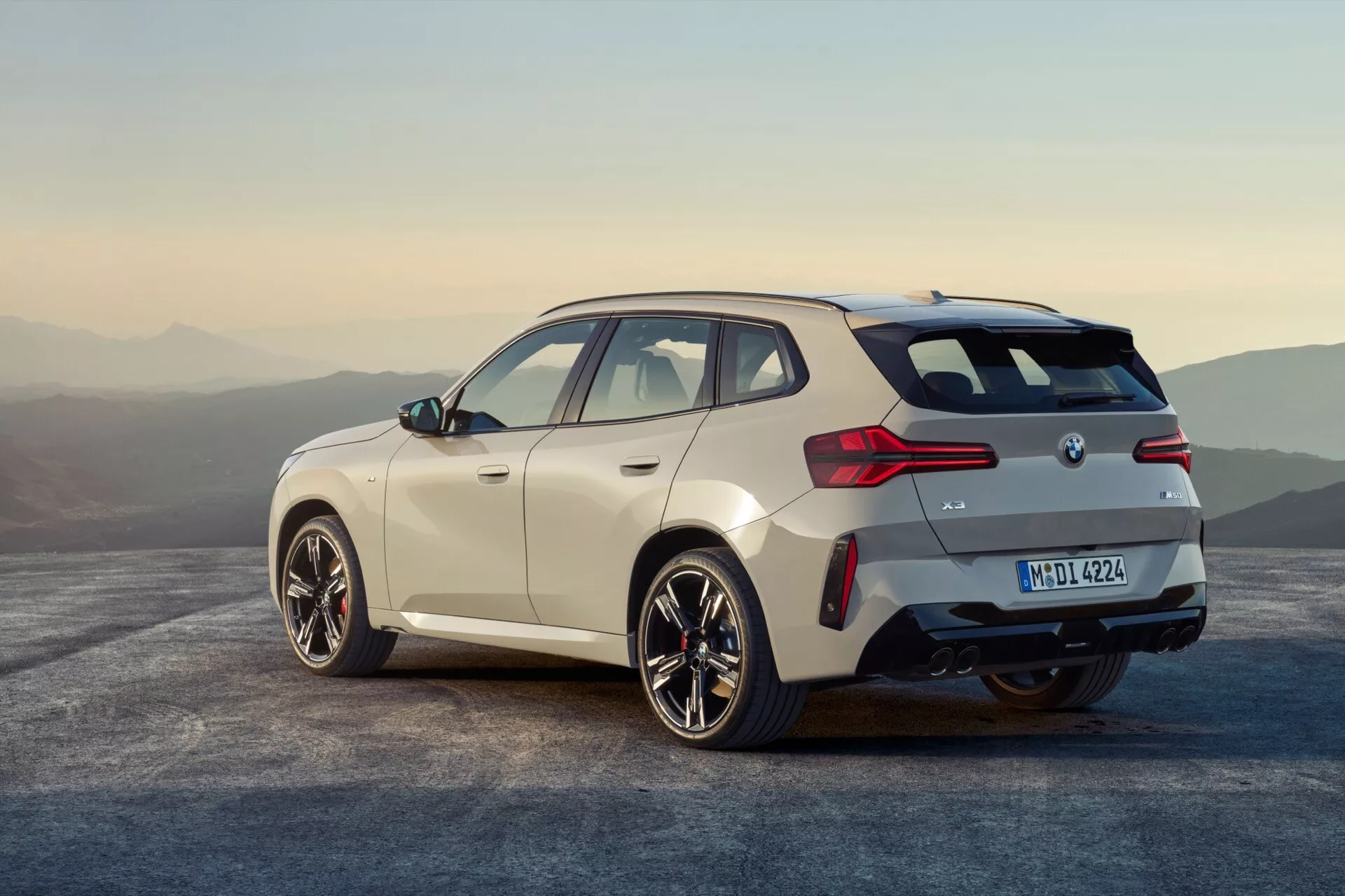 Obraz przedstawia nowoczesny hatchback BMW X3 2024 (G45) z trzech czwartych tyłu, ukazujący opadającą linię dachu z tyłu, jasny błyszczący lakier, duże wieloramienne felgi aluminiowe, tylne światła LED sięgające klapy bagażnika i tylnych błotników, centralnie umieszczone logo marki i tablicę rejestracyjną, dobrze zintegrowany tylny zderzak z widocznymi końcówkami wydechu, eleganckie boczne okna z lusterek z wbudowanymi kierunkowskazami, możliwe relingi dachowe lub spojler oraz naturalne, rozległe tło z mglistymi górami podczas złotej godziny.