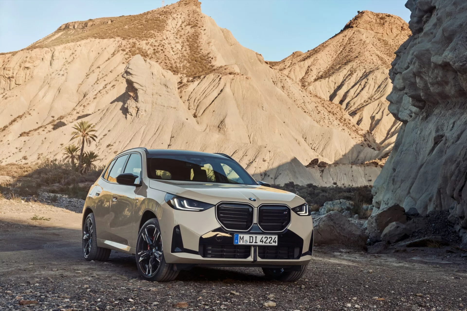Trzy czwarte widoku na BMW X3 (G45) 2024 na krajobrazie pustynnym, z widocznym przednim grillem z charakterystycznym znakiem, smukłymi LED-owymi reflektorami, przednim zderzakiem z wlotami powietrza i sensorami, liniami na masce, wieloramiennymi felgami z lekkich stopów, nisoprofilowymi oponami, bocznymi lusterkami z wbudowanymi kierunkowskazami i jasnym srebrnym kolorem kontrastującym z czarnymi akcentami, co sugeruje luksus i nowoczesną technologię motoryzacyjną.