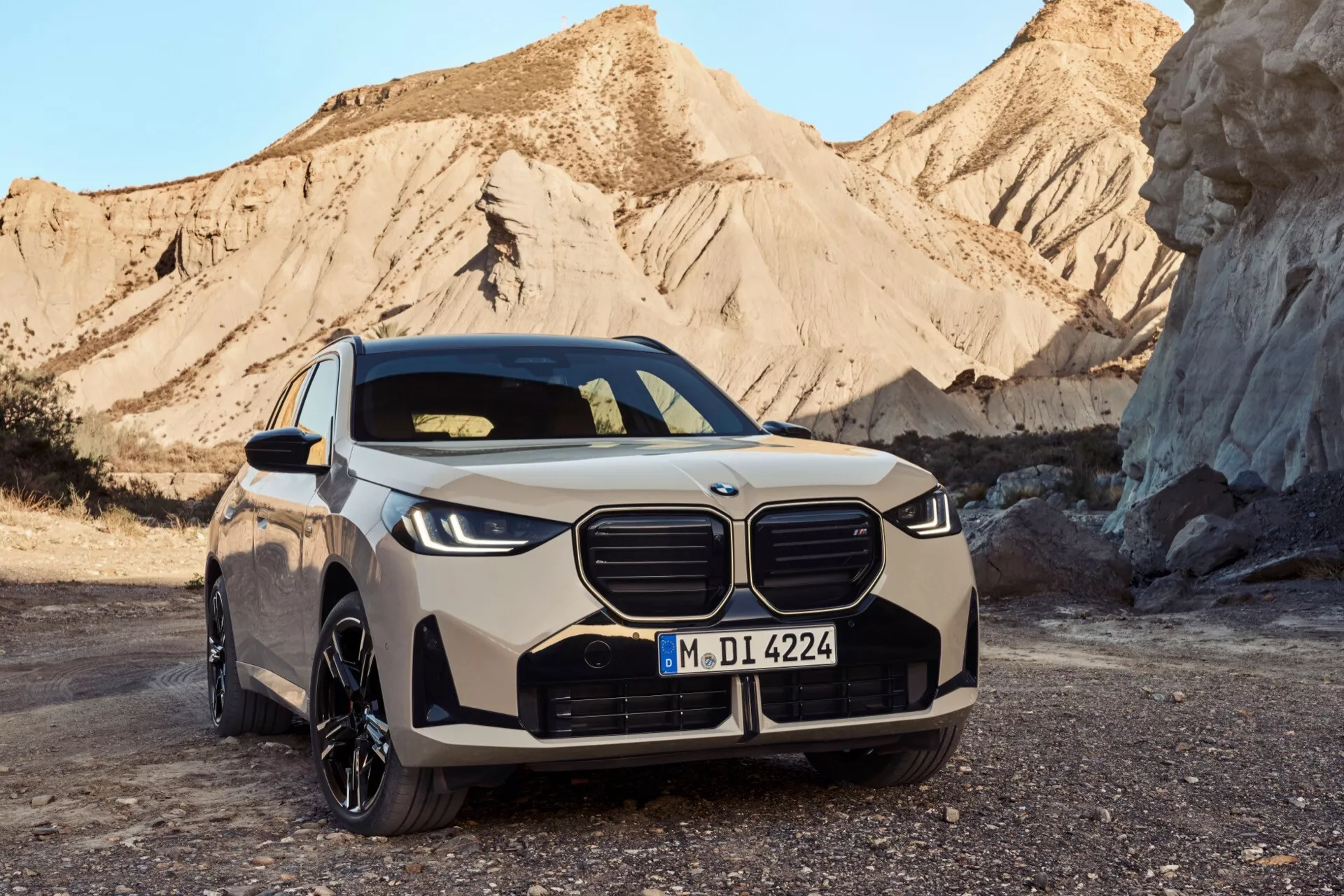 Przedni trzy-czwarte widok BMW X3 2024 (G45), luksusowego elektrycznego SUV-a z zamkniętą kratką nerkową BMW z niebieskimi akcentami, smukłymi reflektorami LED, sportowym przednim zderzakiem, profilowaną maską, poszerzonymi błotnikami z dużymi wieloszprychowymi aluminiowymi felgami, widocznymi lusterkami bocznymi z wbudowanymi kierunkowskazami oraz częściowo widocznym profilem bocznym z płynnymi liniami. Umieszczony na kamienistej powierzchni z pustynnym krajobrazem w tle o świcie lub zmierzchu, podkreślając nowoczesny design i ekologiczną naturę pojazdu.