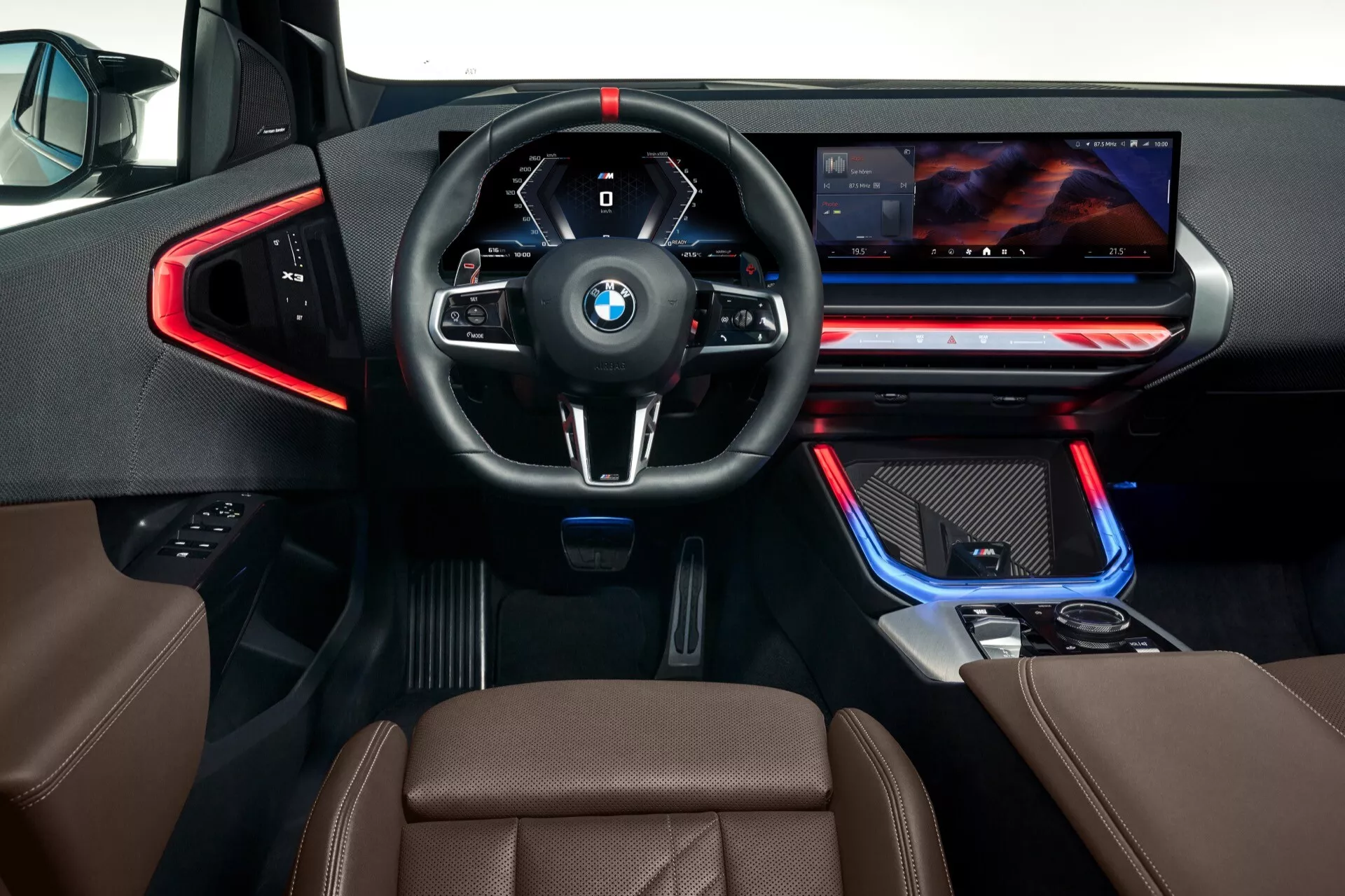 Widok wnętrza BMW X3 2024 (G45) z perspektywy kierowcy. Kąt jest od tyłu, lekko podwyższony nad konsolą środkową, patrząc w dół na deskę rozdzielczą, kierownicę i przednie panele kontrolne. Cechy obejmują skórzaną, wielofunkcyjną kierownicę z czerwonymi szwami, cyfrowy zestaw wskaźników, dźwignię zmiany biegów automatycznej skrzyni biegów, gałkę kontrolną iDrive i szeroki wyświetlacz infotainment. Wielokolorowe oświetlenie LED podkreśla linie wykończeniowe i podnóżki. Przednie siedzenia są tapicerowane brązową skórą. Dodatkowe szczegóły obejmują czerwone akcenty wokół otworów wentylacyjnych i klamek drzwi, metalowe pedały, co sugeruje sportowy wariant.