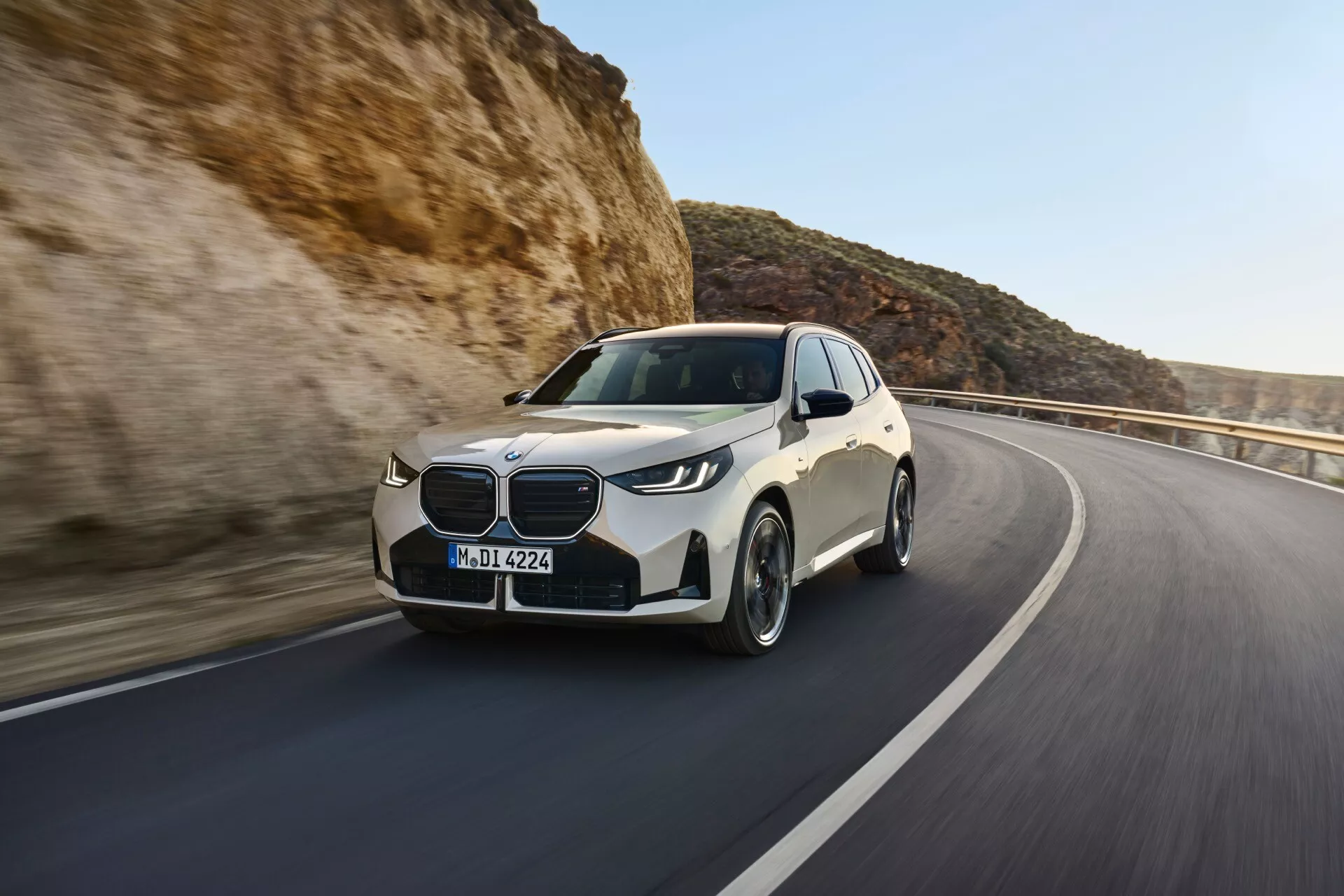 Dynamiczny widok z trzech czwartych przodu BMW X3 2024 (G45) poruszającego się po drodze w pustynnym terenie. Uchwycone nieco od lewej strony, zdjęcie podkreśla stronę kierowcy i przedni grill. Widoczne cechy to elegancki przedni lewy reflektor LED, wyraźny luksusowy grill, rzeźbione linie na masce, wieloramienne felgi sugerujące prędkość i lusterko boczne z wbudowanymi kierunkowskazami. Refleksyjny lakier auta ostro kontrastuje ze skalistym otoczeniem, podkreślając jego nowy, dobrze utrzymany wygląd. Imponująca reprezentacja nowoczesnego designu, wydajności i estetyki samochodu.