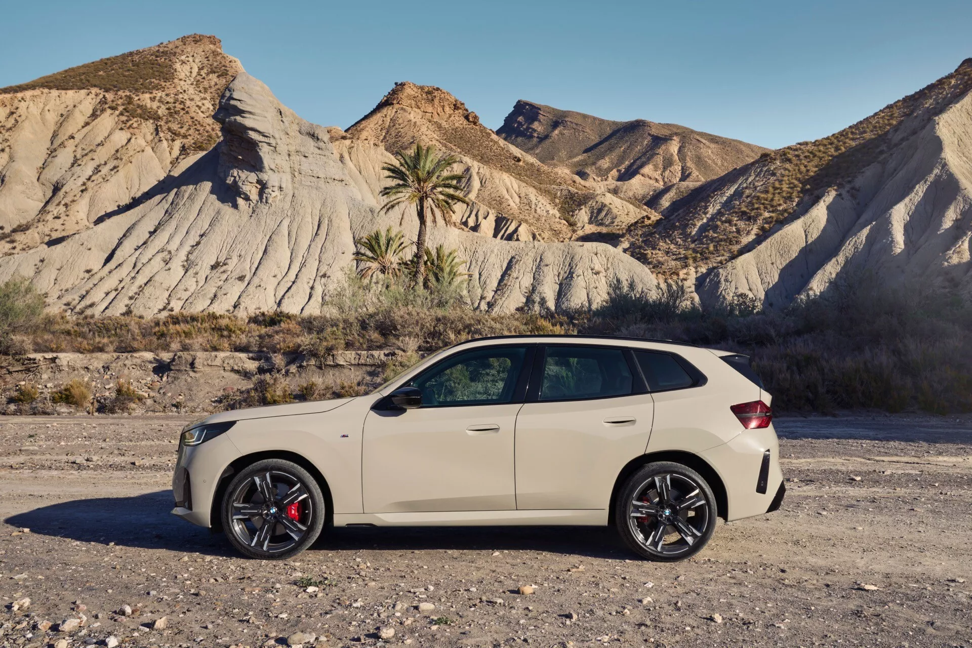 Zdjęcie BMW X3 2024 (G45), nowoczesnego kompaktowego lub średniej wielkości SUV-a z aerodynamicznymi liniami projektowymi, dużymi ciemnymi kołami aluminiowymi z czerwonymi zaciskami hamulcowymi, eleganckimi lusterkami bocznymi z wbudowanymi kierunkowskazami, metalicznym wykończeniem, liniami konturowymi podkreślającymi klamki drzwi, przyciemnianymi szybami i szyberdachem. SUV jest zaparkowany na żwirowej nawierzchni z suchym, pagórkowatym tłem ze skąpą roślinnością, podkreślając jego przygodowy wygląd. Dodatkowe funkcje obejmują relingi dachowe, antenę typu płetwa rekina, tylne światła LED i podwójny układ wydechowy.