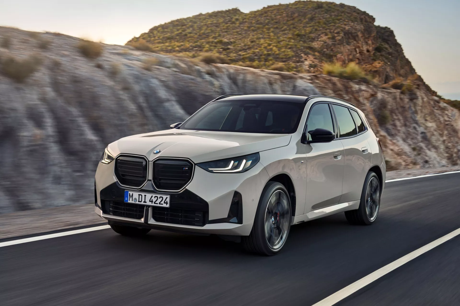 Nowoczesny luksusowy crossover SUV BMW X3 2024 (G45) uchwycony w ruchu pod kątem trzech czwartych przodu, ukazujący przedni grill, zestaw świateł, przedni zderzak, maskę, błotnik po stronie kierowcy i koła. Usytuowany na zakręcie drogi z rozmytym tłem sugerującym prędkość i ruch, samochód posiada eleganckie reflektory LED, aerodynamiczne kontury i duże koła, sugerując efektywny design i zaawansowaną technologię.