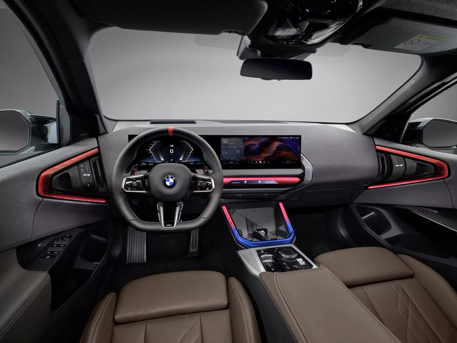 Zdjęcie wnętrza BMW X3 2024 (G45) z perspektywy kierowcy, wykonane z centralnego tylnego kąta na wysokości tylnego siedzenia. Na pierwszym planie skórzana kierownica z wielofunkcyjnymi przyciskami i cyfrowym zestawem wskaźników. Konsola centralna z dźwignią zmiany biegów, kontrolą trybów jazdy oraz prawdopodobnie interaktywnym touchpadem. Szeroki ekran infotainment w desce rozdzielczej na jednym poziomie. Oświetlenie ambientowe wzdłuż deski rozdzielczej i drzwi. Wnętrze z luksusowymi, skórzanymi siedzeniami, wysokiej jakości wykończeniami i sportowymi fotelami przednimi z wyraźnie zarysowanymi bokami, sugerujące luksus i orientację na osiągi. Auto wydaje się być w stanie postoju, dobrze oświetlone, bez osób w zasięgu wzroku.
