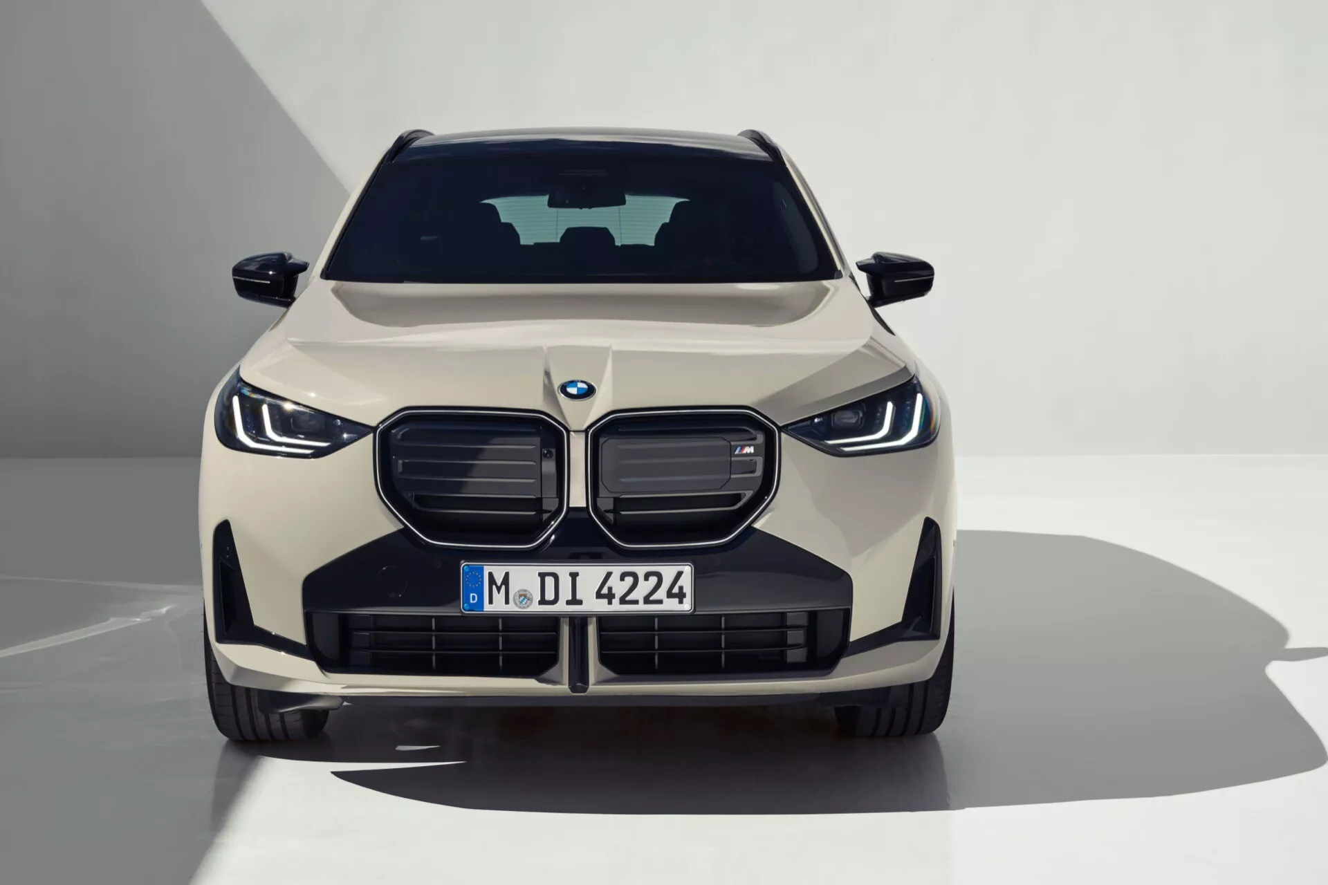 Przód, symetryczny obraz BMW X3 2024 (G45) oświetlony w studio, ukazujący dużą, solidną kratkę z emblematem, eleganckie reflektory LED, czujniki zaawansowanych systemów wspomagania kierowcy, konturowane linie maski i błyszczące wykończenie w kolorze jasnobeżowym lub przytłumionym złotym