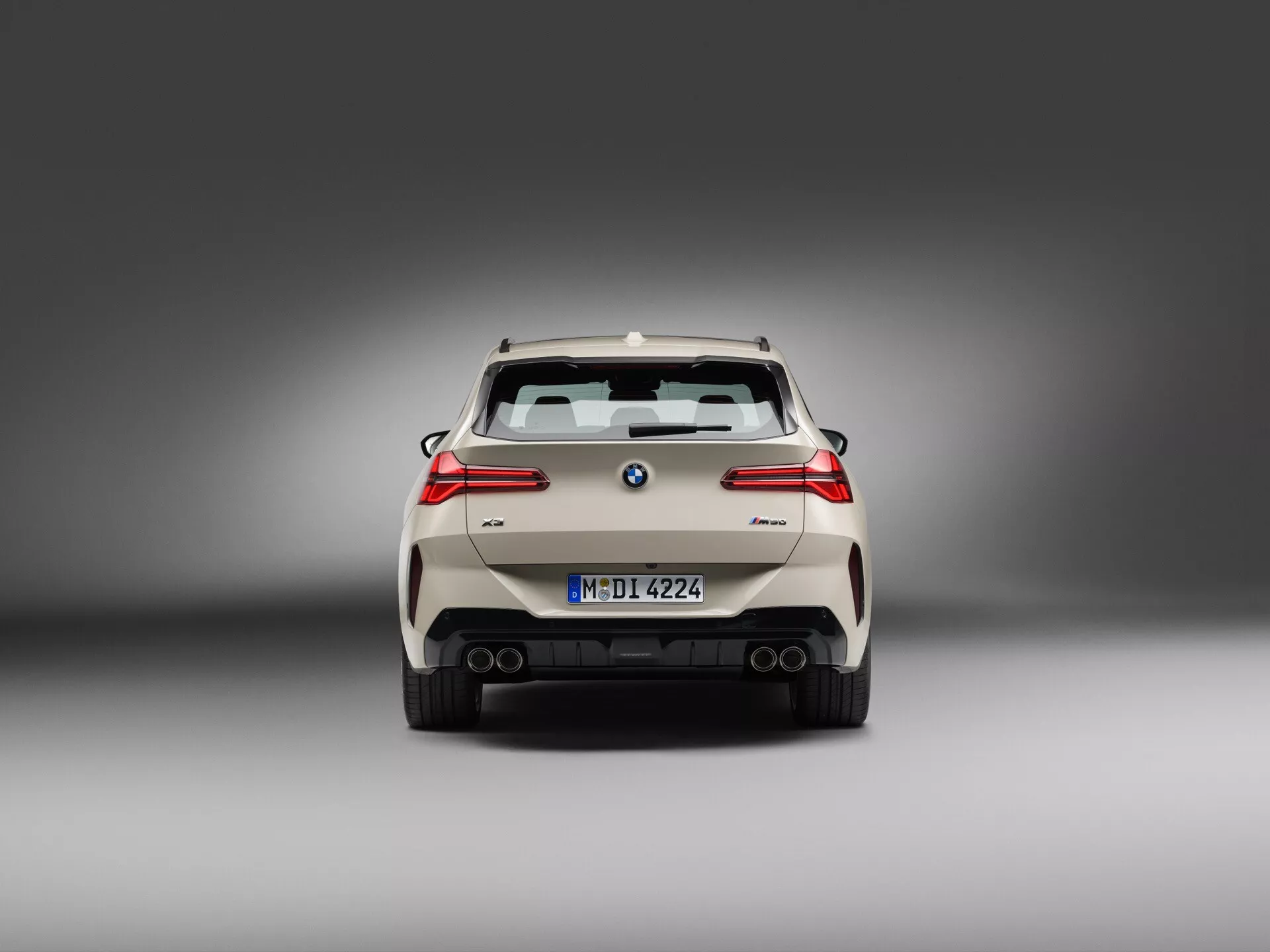Profesjonalne zdjęcie studyjne, widok tylny BMW X3 2024 (G45) ukazujące charakterystyczne elementy designu. Samochód jest wyśrodkowany, z wyraźną tablicą rejestracyjną poniżej logo producenta, otoczony wygaszonymi tylnymi światłami. Widoczne są reflektory na zewnętrznych krawędziach zderzaka, podwójny układ wydechowy, tylna szyba z wycieraczką i spojler dachowy. Zderzak posiada czujniki parkowania oraz dyfuzor, co nadaje sportowy wygląd. Miękkie oświetlenie ujawnia częściowo widoczne wieloramienne felgi, umieszczone na neutralnym szarym tle gradientowym.
