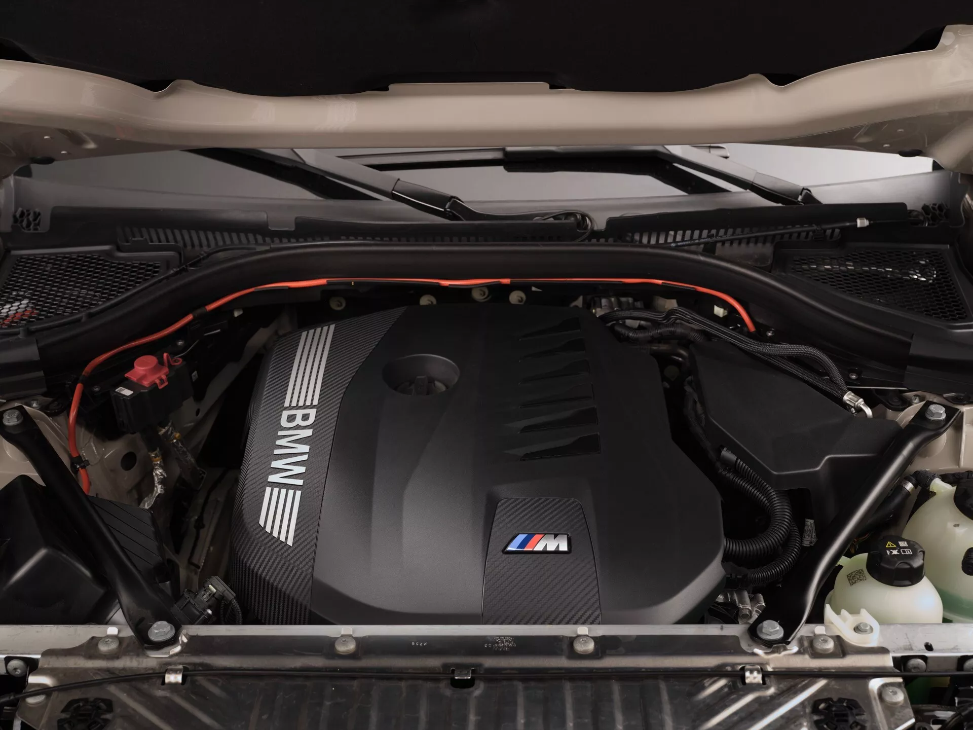 Zbliżenie komory silnika BMW X3 2024 (G45) z perspektywy z góry z podniesioną maską. Pokrywa silnika z napisem 'BMW M' wykonana jest z tworzywa sztucznego wzmacnianego włóknem węglowym, ma błyszczącą, ciemną kolorystykę i ozdobiona jest paskami logo BMW M. Widoczne są różne komponenty silnika, takie jak izolacyjne wykładziny, części nadwozia z matowym wykończeniem, elastyczne czerwone akcenty, pojemniki na płyny, kable i węże, podkreślające czystą i starannie zagospodarowaną komorę silnika.