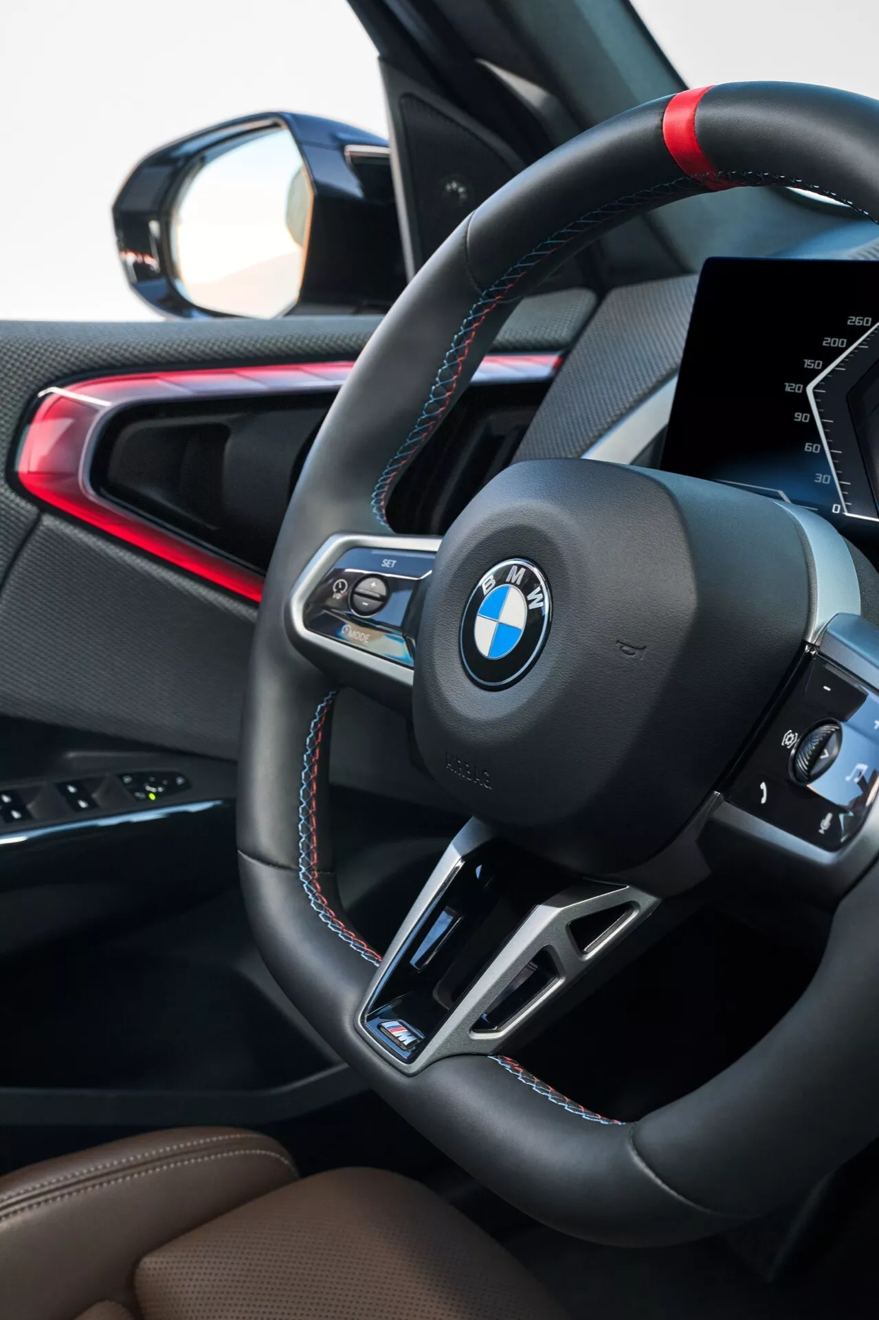 Zbliżenie wnętrza BMW X3 2024 (G45) skupione na kierownicy i miejscu kierowcy. Sportowa kierownica obszyta skórą z kontrastowym czerwono-niebieskim ściegiem z serii M Performance BMW ma wielofunkcyjne przyciski, łopatki do zmiany biegów oraz logo BMW. Widoczny jest częściowy cyfrowy wyświetlacz, wskazujący na nowoczesną technologię. Dodatkowe detale obejmują brązowe skórzane siedzenia oraz czerwoną linię akcentującą na drzwiach pasażera lub konsoli środkowej.