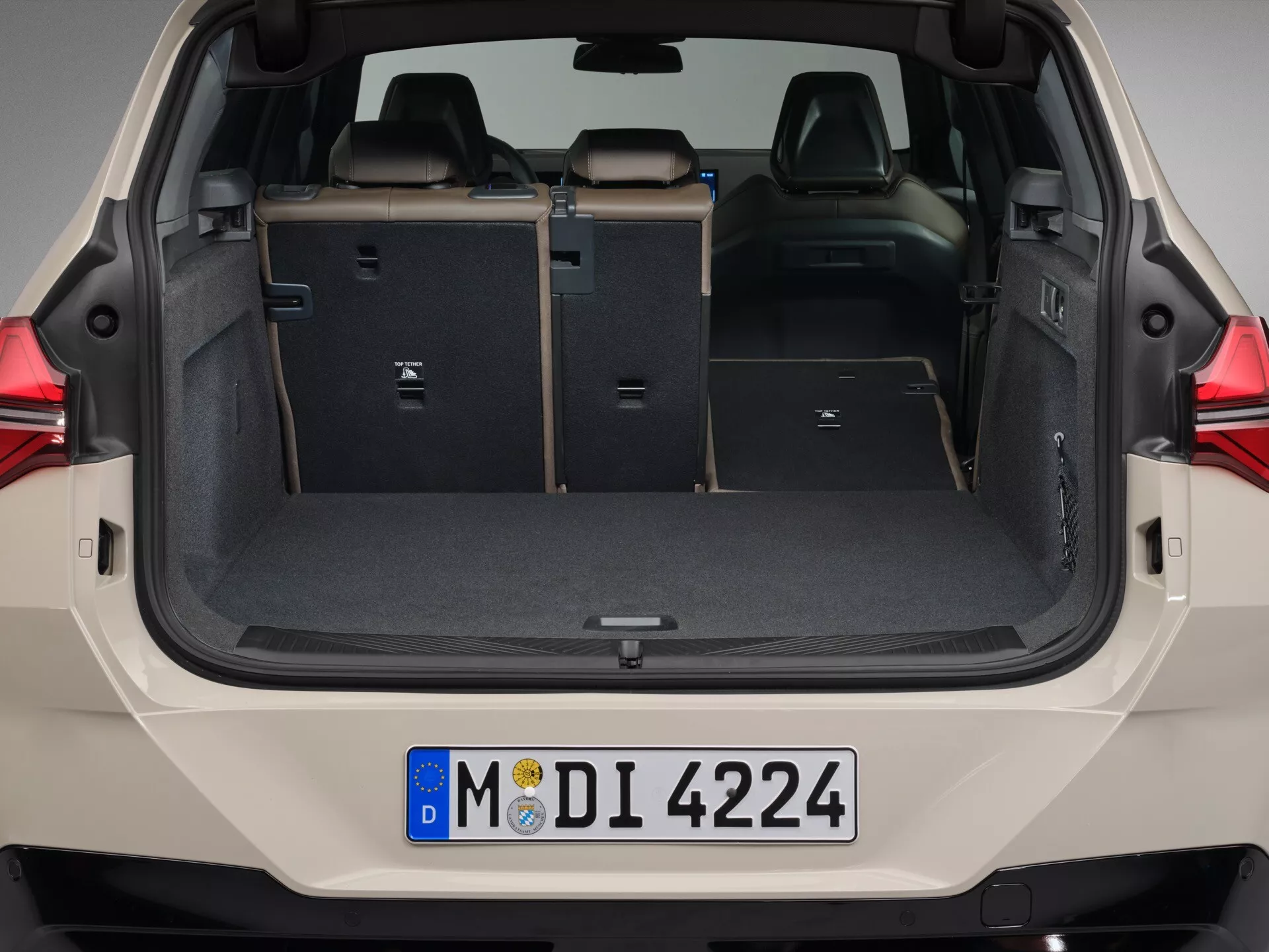 Obraz przedstawiający przestrzeń ładunkową BMW X3 2024 (G45) z widoku tylnego, z całkowicie otwartą klapą. Zdjęcie na poziomie oczu ukazuje czystą, pustą przestrzeń bagażnika z podniesionymi tylnymi siedzeniami, posiadającymi widoczne mechanizmy zatrzaskowe i funkcję dzielonego składania dla elastycznego przewozu ładunku i pasażerów. Wykończenie wnętrza tapicerowane, z płaską, wysuwaną osłoną na bagażnik dla prywatności i ochrony. Mniejsze przegrody i haczyki po wewnętrznych stronach służą do celów organizacyjnych. Nowoczesny design z czystymi liniami i wysokiej jakości materiałami sugeruje dobrze wyposażony pojazd, prawdopodobnie SUV lub hatchback, wskazywany przez częściowo widoczne tylne światła i europejską tablicę rejestracyjną.