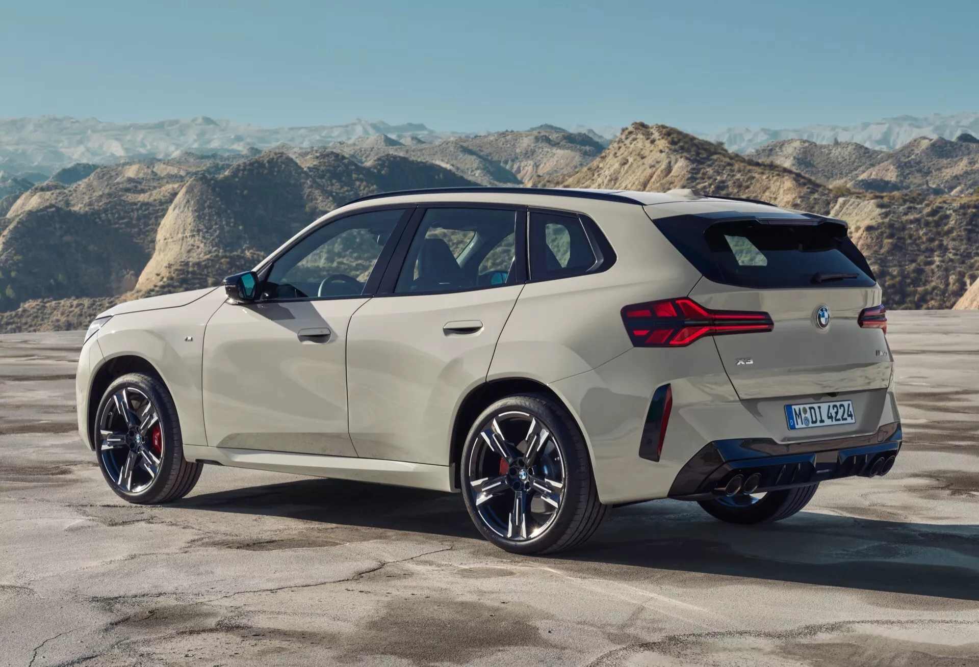 Zdjęcie BMW X3 2024 (G45) z perspektywy tylniej w trzech czwartych, podkreślające jego elegancki, aerodynamiczny profil, skomplikowane wieloszprychowe felgi aluminiowe, wyraźną linię charakterystyczną biegnącą pod oknami, sportowe boczne szyby zwężające się ku tyłowi, nowoczesne tylne klosze świetlne LED, podwójny układ wydechowy i prawdopodobne funkcje asysty parkowania w tylnym zderzaku. Pojazd prezentuje luksusową, sportową estetykę z ewentualnym napędem hybrydowym.