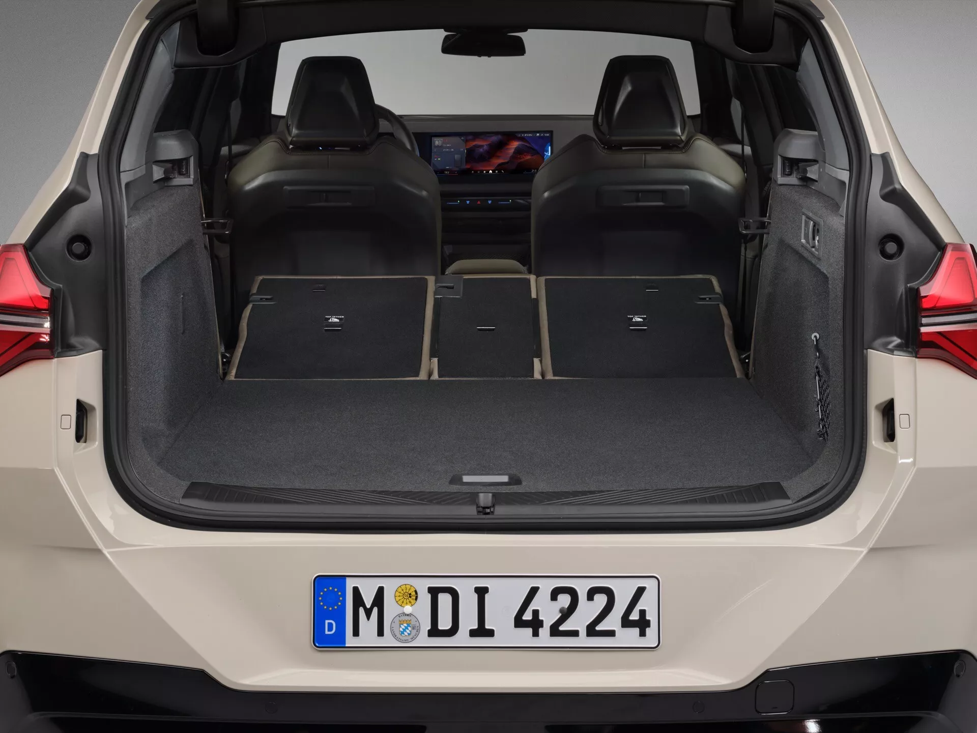 Zdjęcie przedstawia tylne miejsce ładunkowe BMW X3 (G45) 2024 z otwartą klapą bagażnika, widziane z nieco wyższego poziomu oczu. Przedstawia pusty bagażnik z rozłożonymi siedzeniami, ujawniając konfigurację tylnej kanapy dzieloną w proporcji 60/40, wnętrze wyłożone czarną wykładziną, plastikowe listwy, haczyki i wnęki do organizacji. Wewnętrzna strona klapy bagażnika ma wcięcie ułatwiające chwytanie, a na tylnym zderzaku widoczna jest niemiecka tablica rejestracyjna. Tył samochodu jest głównie biały z widocznymi tylnymi światłami.