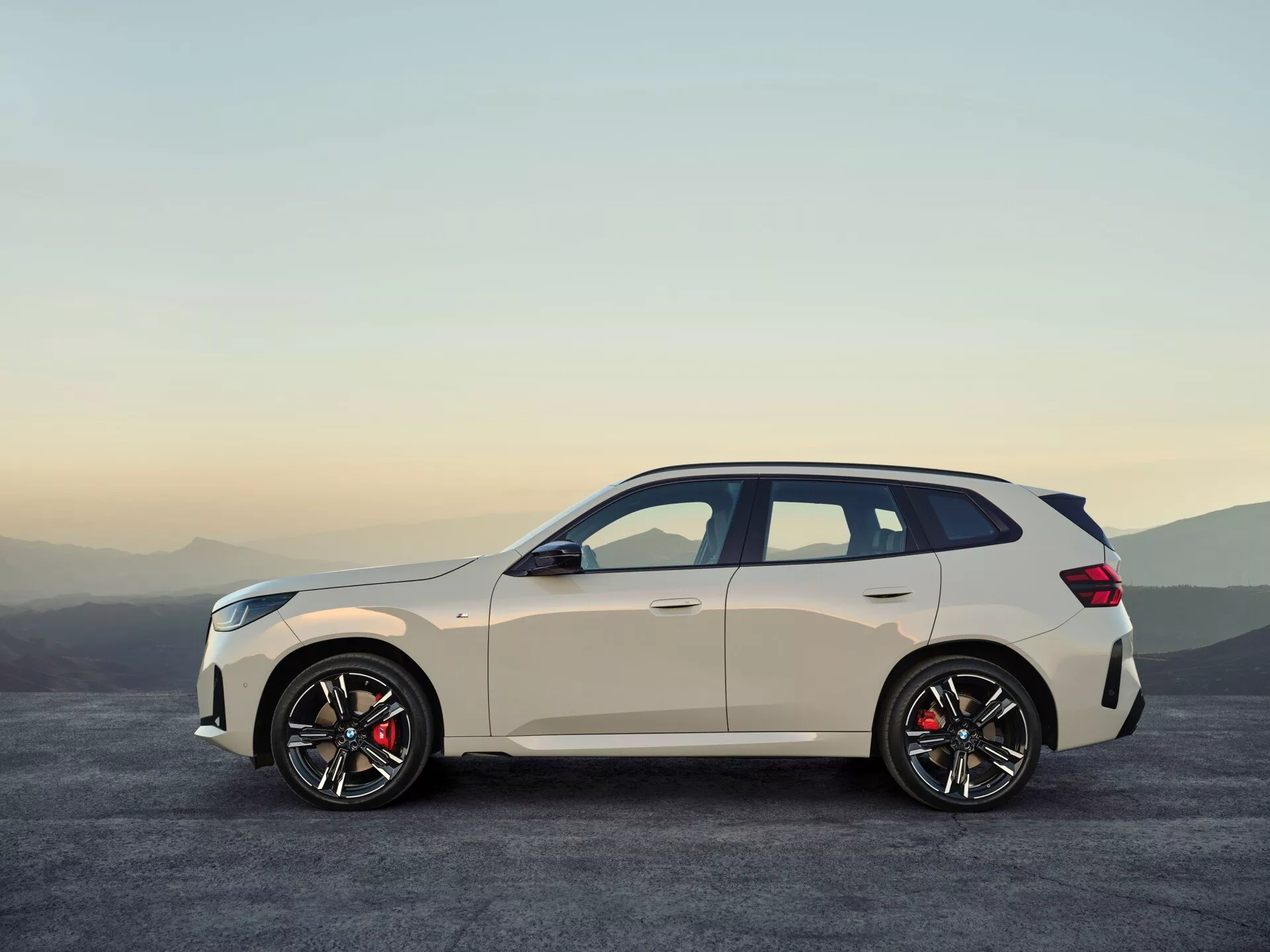 Widok boczny BMW X3 2024 (G45) uchwyconego podczas wschodu lub zachodu słońca. Eleganckie SUV z czterem drzwiami, o jasnej karoserii kontrastującej z ciemnymi oknami i kołami, prezentuje sportowy design. Posiada delikatnie opadającą linię dachu, duże koła wieloszprychowe z czerwonymi zaciskami hamulcowymi, boczne progi i wpuszczane klamki. Ustawiony na tle spokojnego naturalnego krajobrazu na równą nawierzchnię asfaltową, obraz wyróżnia dynamiczne i premium cechy pojazdu.