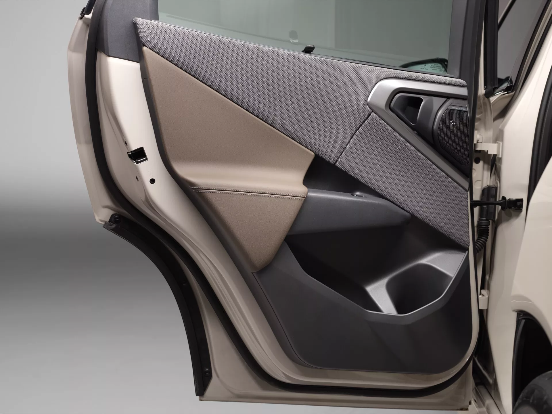 Obraz pokazuje zbliżenie panelu wewnętrznego drzwi BMW X3 2024 (G45). Drzwi mają dwukolorowe wykończenie z gładkim jasnobeżowym lub kremowym kolorem na górze i ciemniejszym kolorem, prawdopodobnie czarnym lub ciemnoszarym, na dole. Materiały wydają się być mieszanką wysokiej jakości skór lub materiałów podobnych do skóry i tkanin. Kątownik w ciemniejszym odcieniu nadaje nowoczesny wygląd. Solidna, wytrzymała klamka ma matowe wykończenie, a tuż poniżej widoczny jest panel sterowania do elektrycznych szyb i prawdopodobnie zamków, co podkreśla ergonomiczną konstrukcję. Przestronny schowek jest wbudowany w dolny panel. Zdjęcie jest zrobione pod lekko podwyższonym, skośnym kątem, skupiając się na środkowej części drzwi, z miękkim, rozproszonym oświetleniem podkreślającym szczegóły.