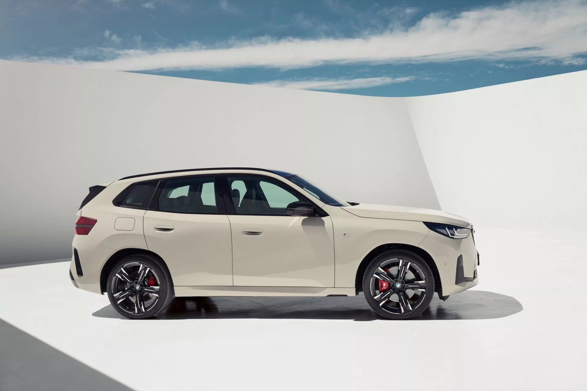 Zdjęcie białego BMW X3 2024 (G45) kombi, uchwyconego z bocznego profilu, skupiającego się na stronie pasażera. Samochód jest ustawiony w studiu z neutralnym tłem, podkreślając jego luksusowy design z cechami takimi jak ostre linie nadwozia, eleganckie klamki, aerodynamiczne lusterka boczne i duże felgi aluminiowe z czerwonymi zaciskami hamulcowymi. Pojazd jest zaparkowany z prostymi kołami, pokazując swój współczesny styl bez włączonych świateł.