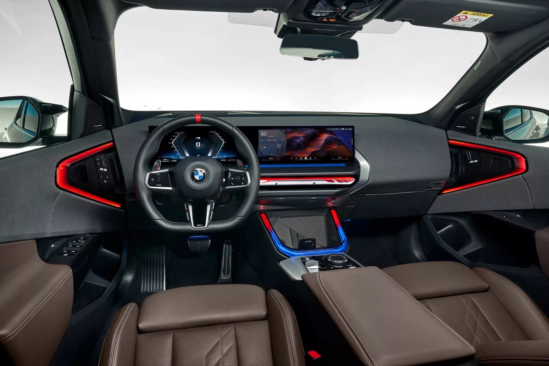 Obraz przedstawia wnętrze BMW X3 2024 (G45) z poziomu oczu, skoncentrowane na desce rozdzielczej i sterowaniu kierowcy. Wielofunkcyjna, trójramienna kierownica zawiera logo BMW i przyciski sterujące po obu stronach. Za nią znajduje się duży, cyfrowy panel przyrządów, który dostarcza kluczowe informacje o pojeździe na szerokim ekranie. Centralny wyświetlacz infotainmentowy rozciąga się w kierunku pasażera, otoczony minimalistycznymi kratkami wentylacyjnymi i eleganckim panelem kontrolnym dla klimatyzacji i innych funkcji. Konsola centralna zawiera elektroniczną dźwignię zmiany biegów, ambientowe oświetlenie i pokrętło sterujące podobne do iDrive. Tapicerka przednich siedzeń z brązowej lub beżowej skóry dodaje luksusowego akcentu. Zdjęcie podkreśla nowoczesne, wysoko zaawansowane technologicznie wnętrze, ze szczególnym skupieniem się na obszarze kierowcy.